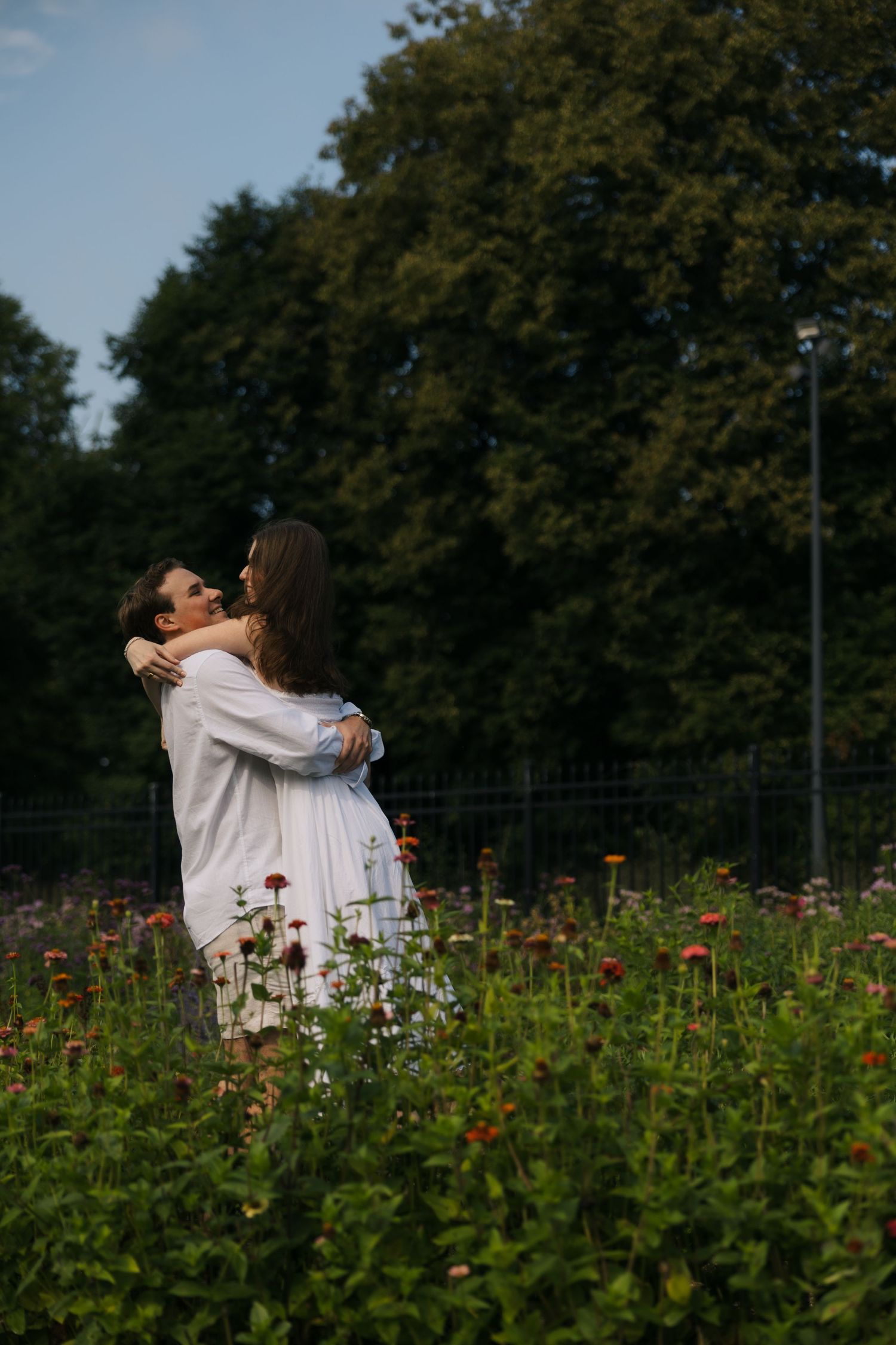 Couple hugging in a colorful flower field with tall green trees and blue sky in the background.