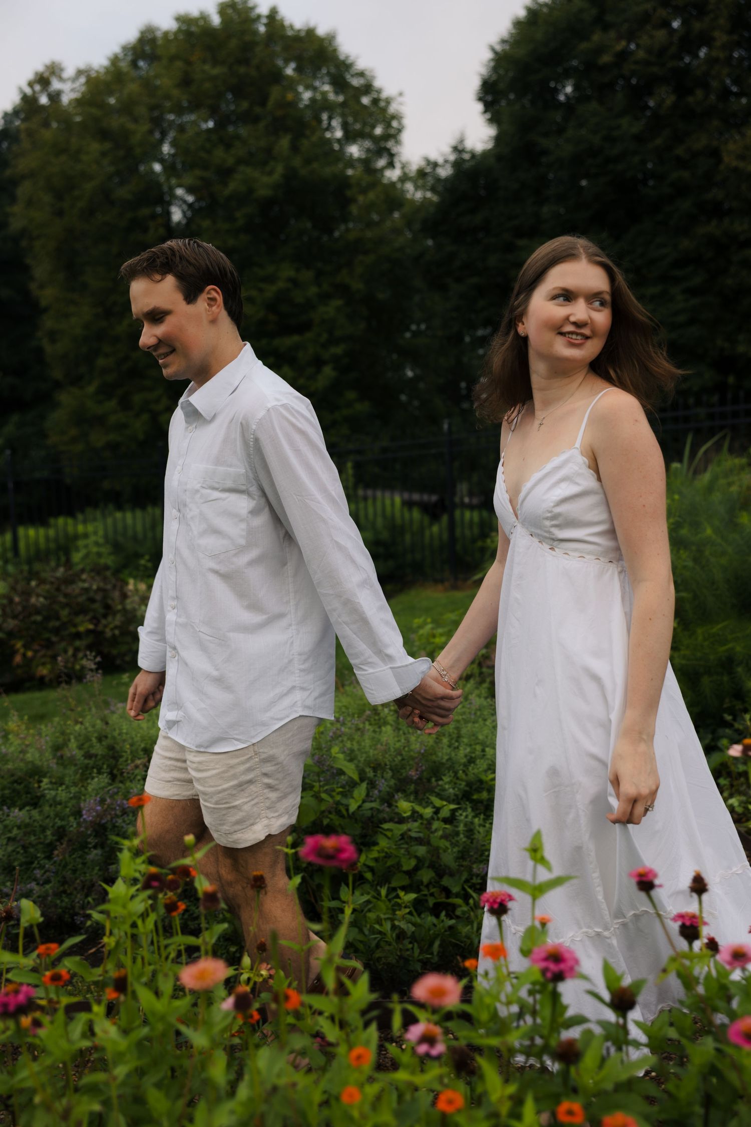 A couple in white outfits holds hands while walking through a lush garden with colorful flowers and green trees.