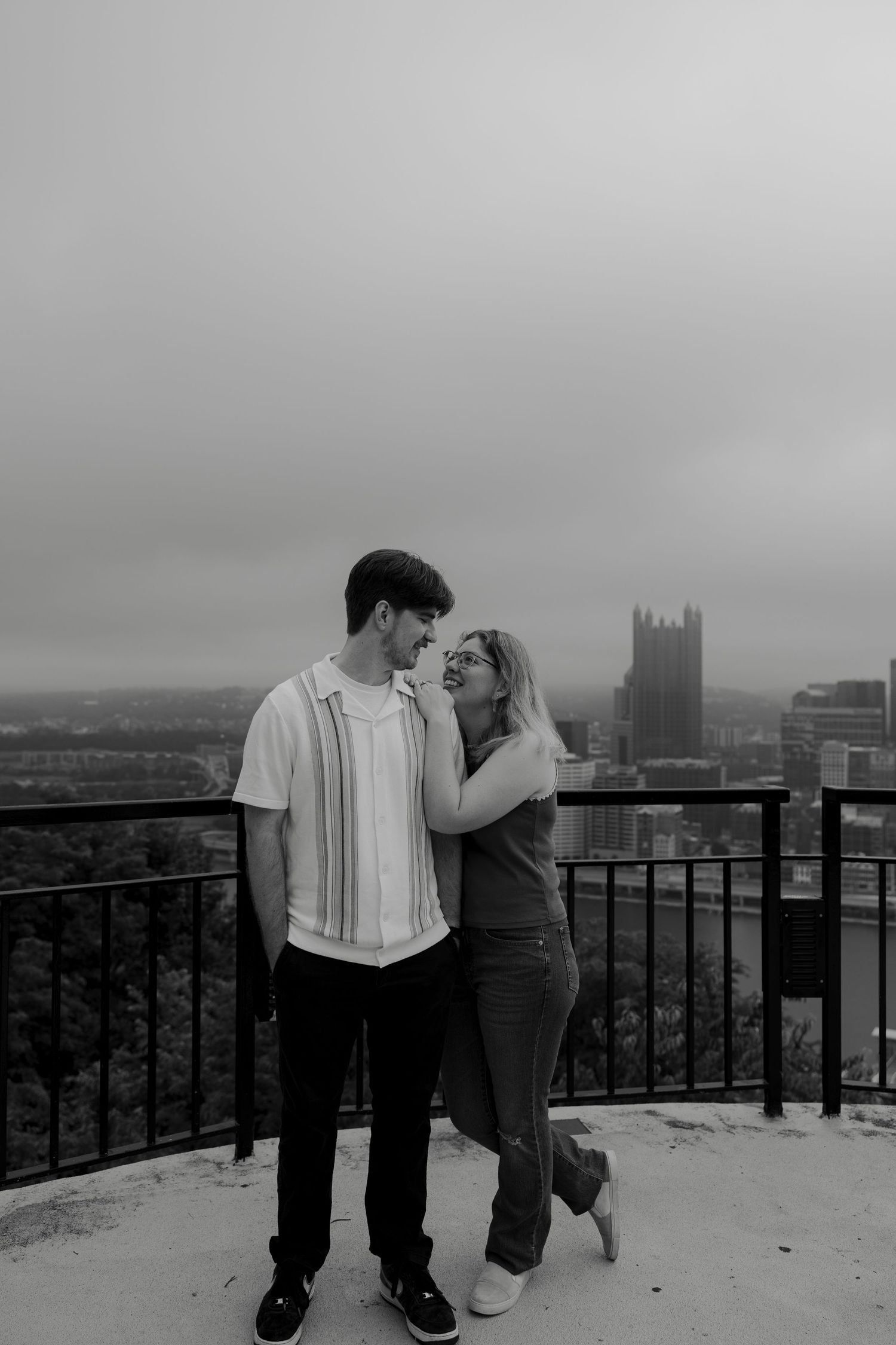 Black and white photo of a couple sharing a romantic moment on a rooftop overlook with Pittsburgh skyline behind them.
