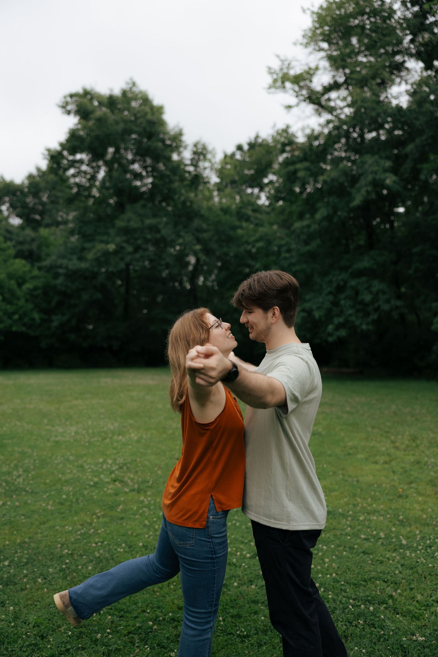 Happy couple dancing together on a green lawn, holding hands and smiling, with tall trees in the background.