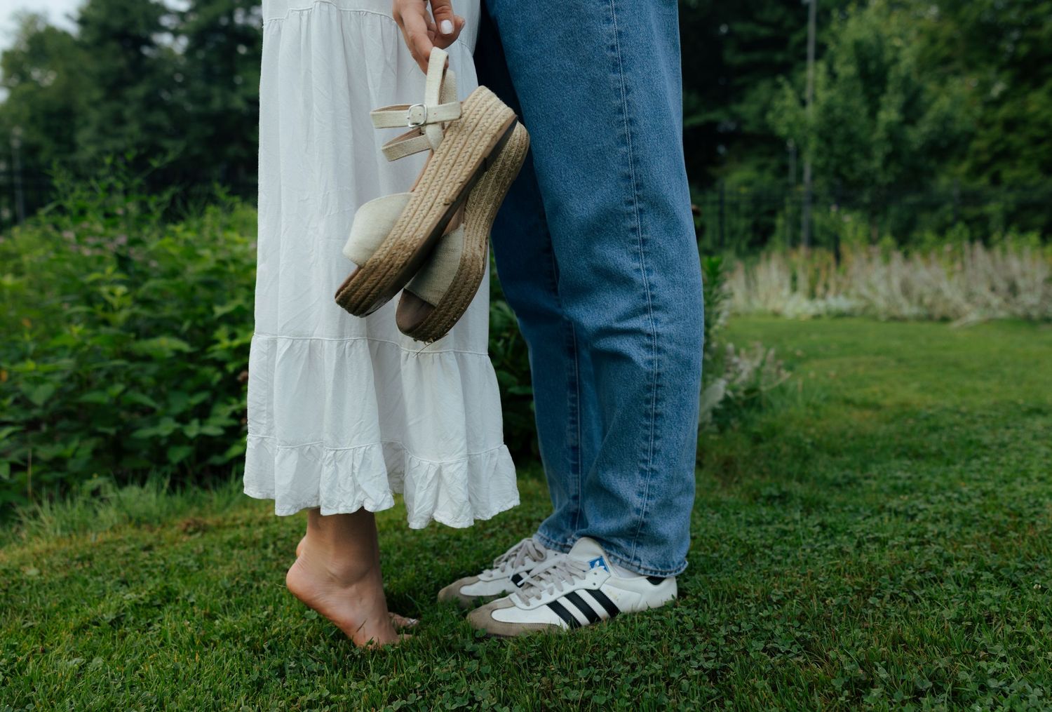 A barefoot woman in a white dress holds her platform shoes beside a man wearing Adidas sneakers on grass.
