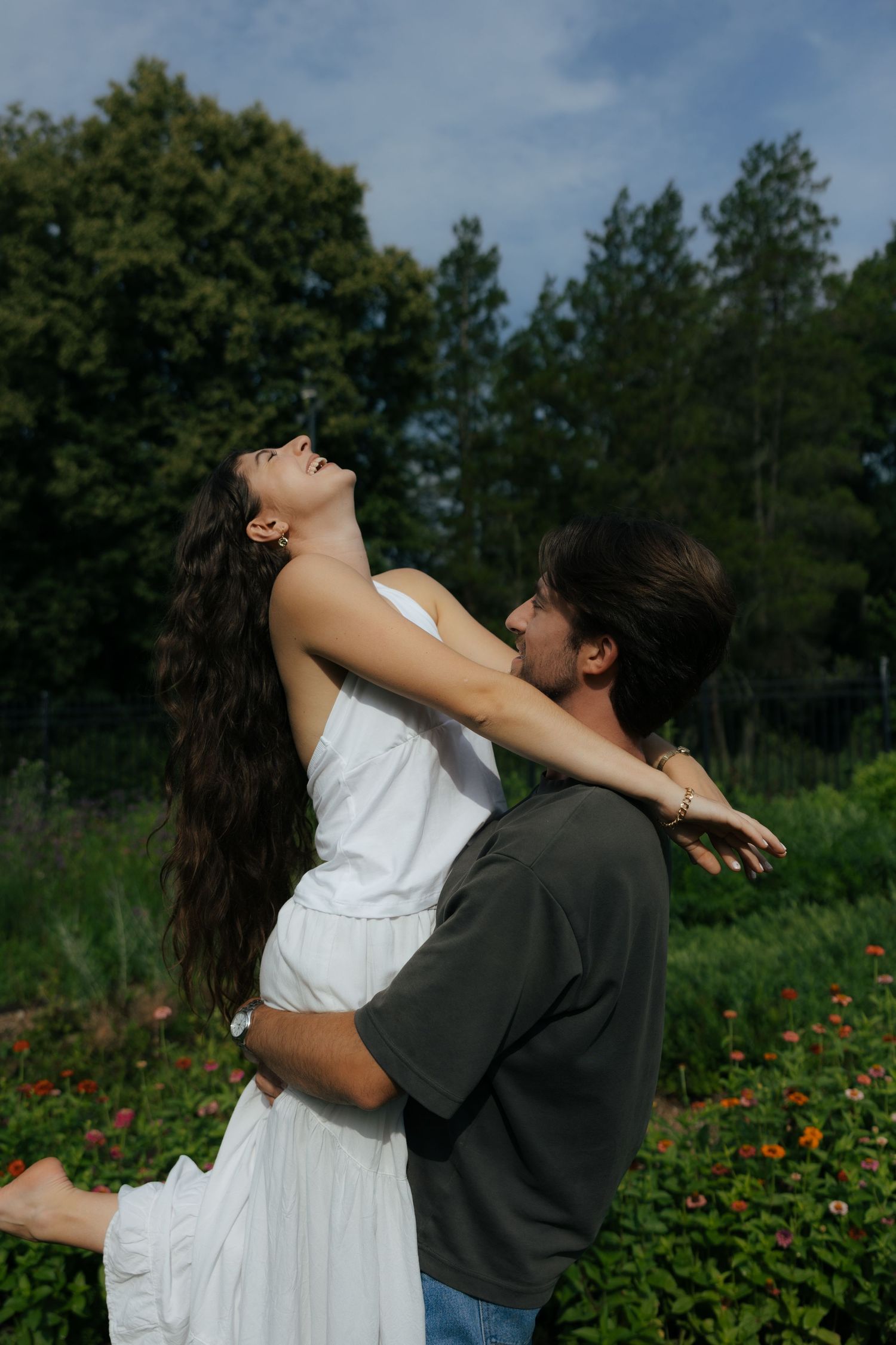 Man holding woman in white dress as she leans back laughing joyfully in a vibrant outdoor garden setting.