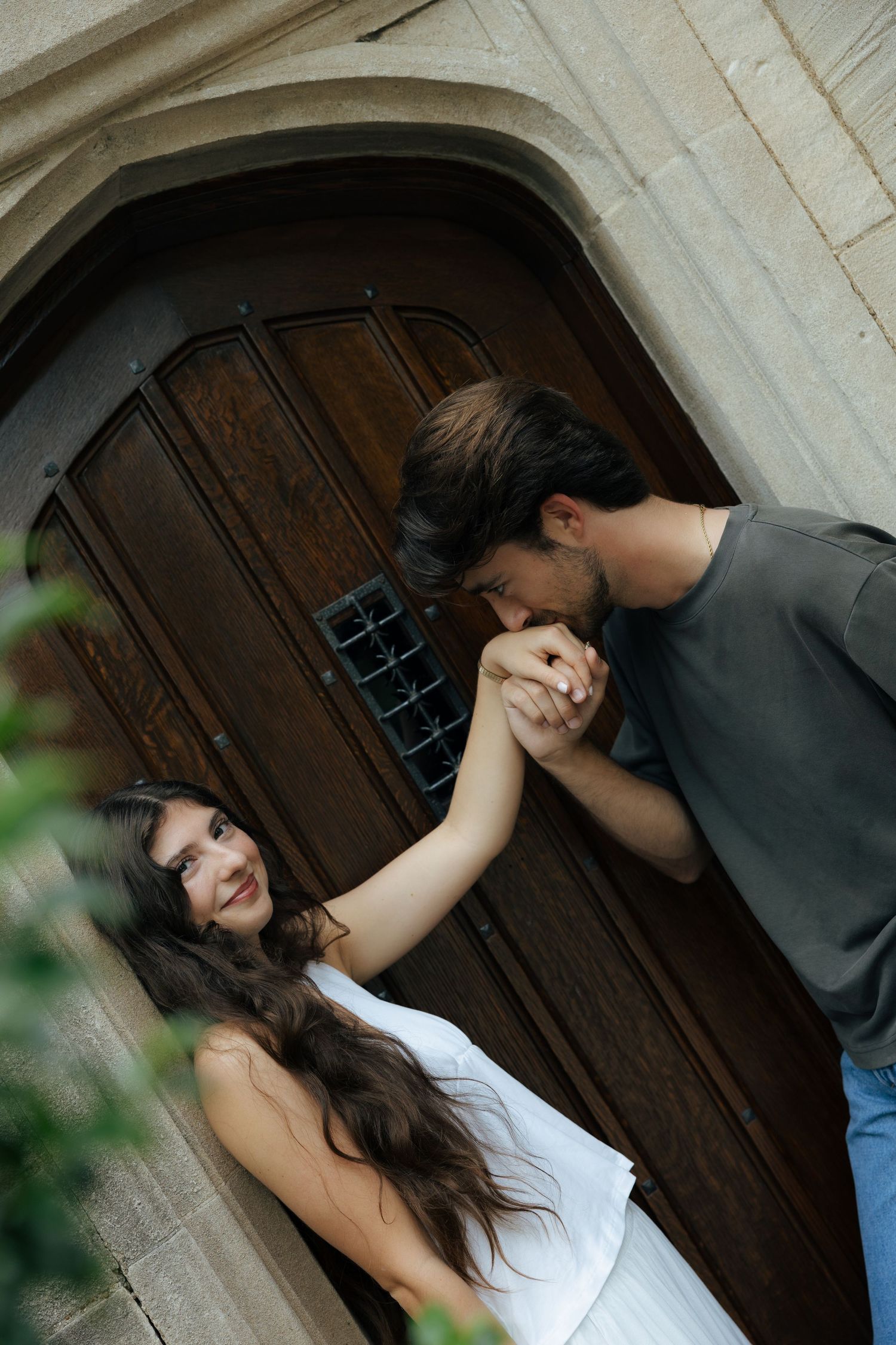 Couple holding hands near vintage wooden arched doorway, man kissing woman's hand tenderly.