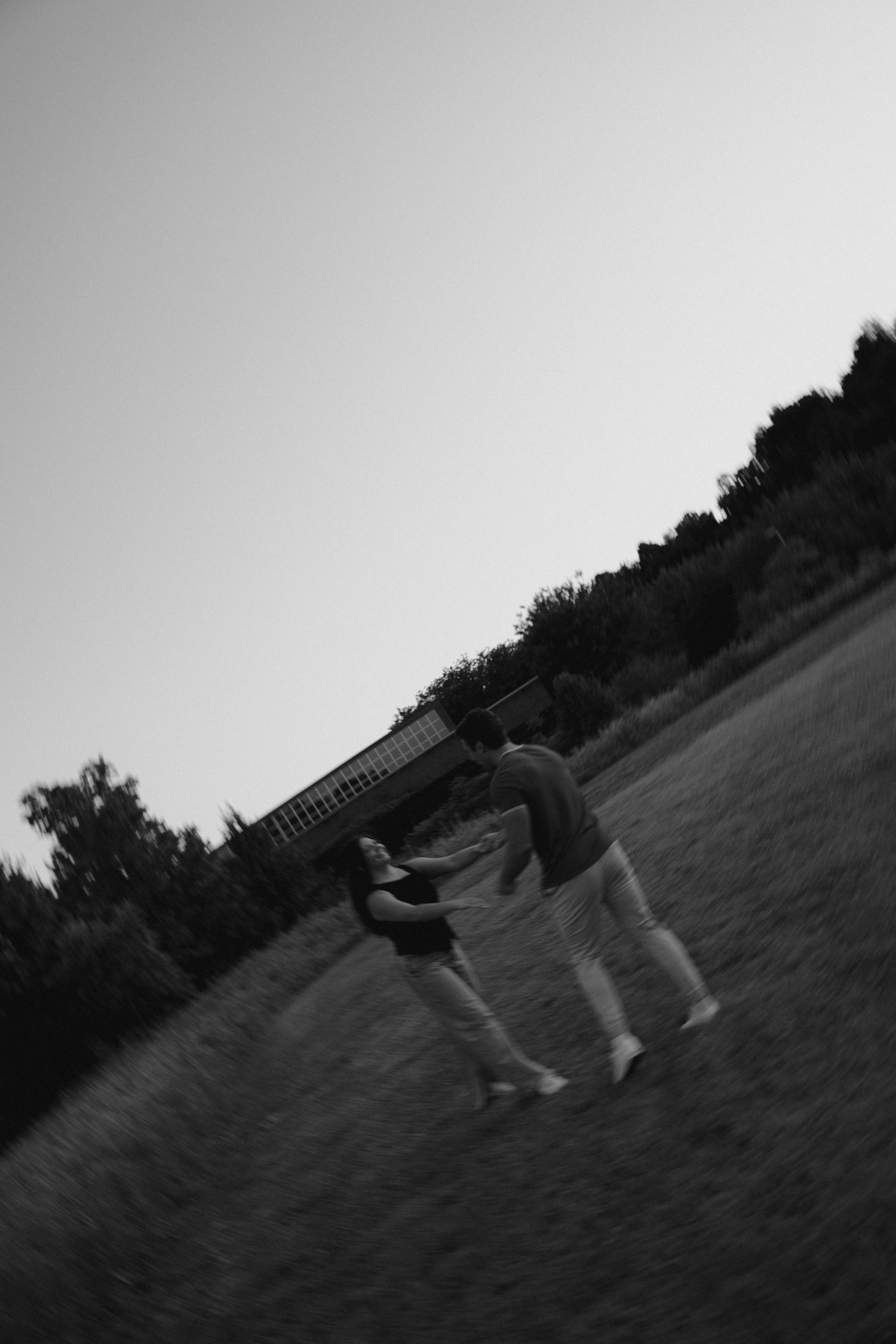 A black and white photo of a couple dancing playfully in an open field at dusk.