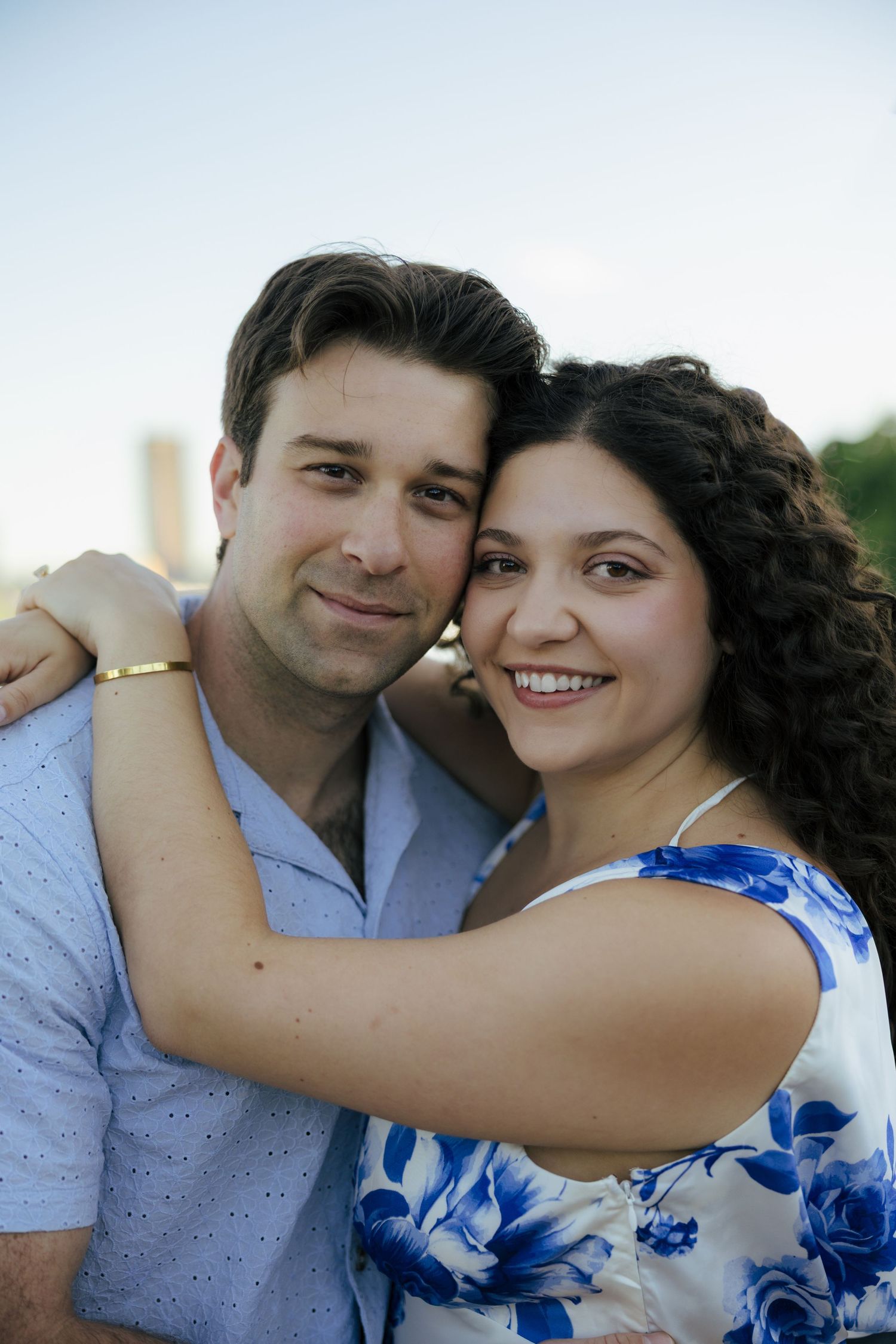 Smiling couple embracing outdoors, woman in blue floral dress and man in light blue shirt, with city skyline in background.