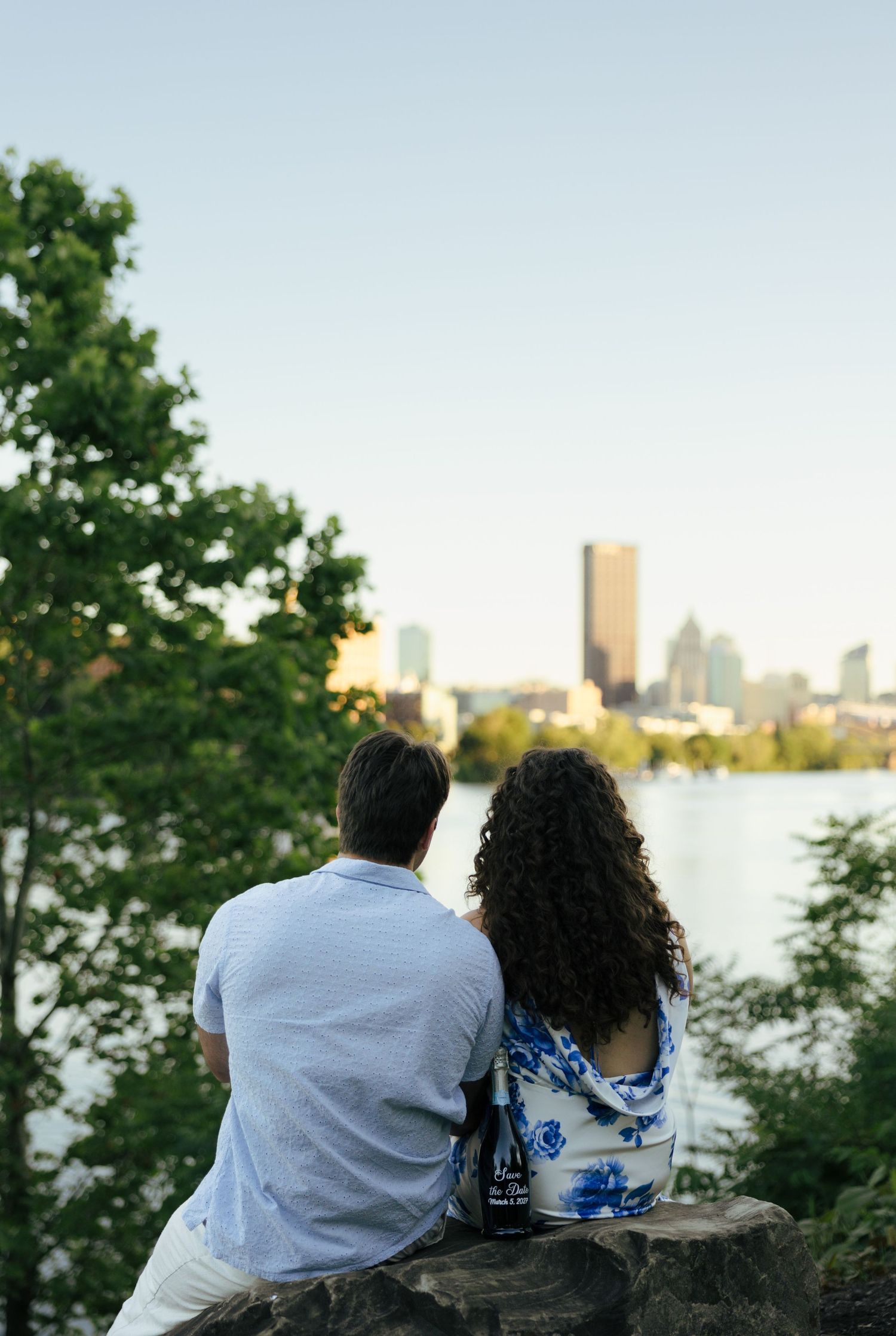 An engaged couple sits on a rock overlooking a city skyline and river at golden hour.