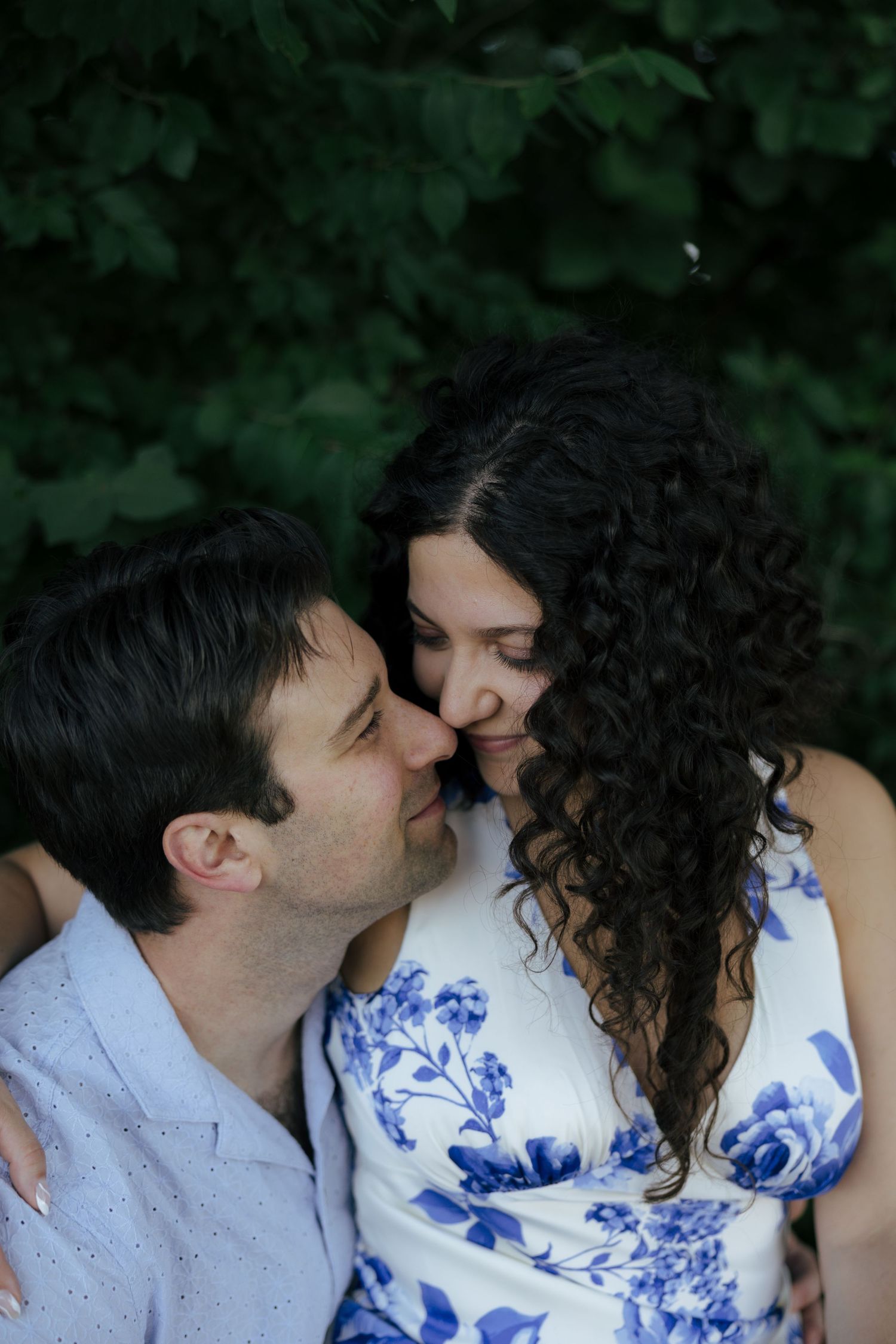 Couple sharing an intimate nose-to-nose moment while seated against a dark green leafy backdrop.