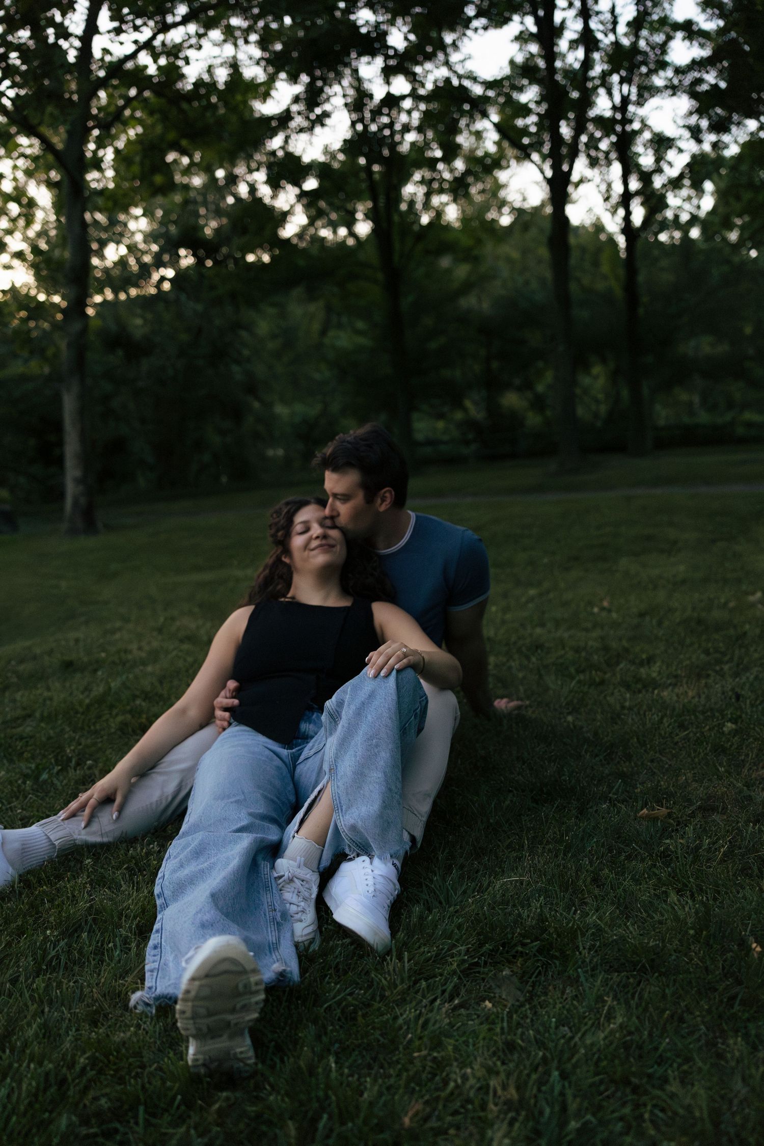 Couple sitting on grass at dusk, man kissing womans head while she leans back on him.