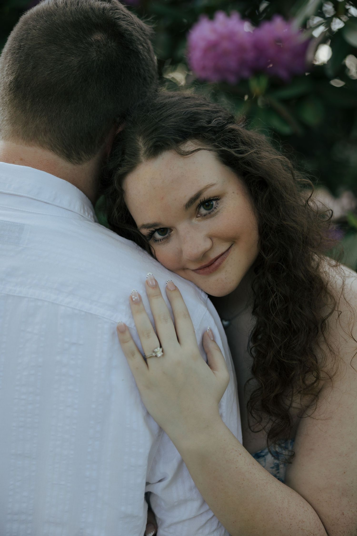 Woman with curly hair smiles over mans shoulder showing engagement ring with purple flowers behind.