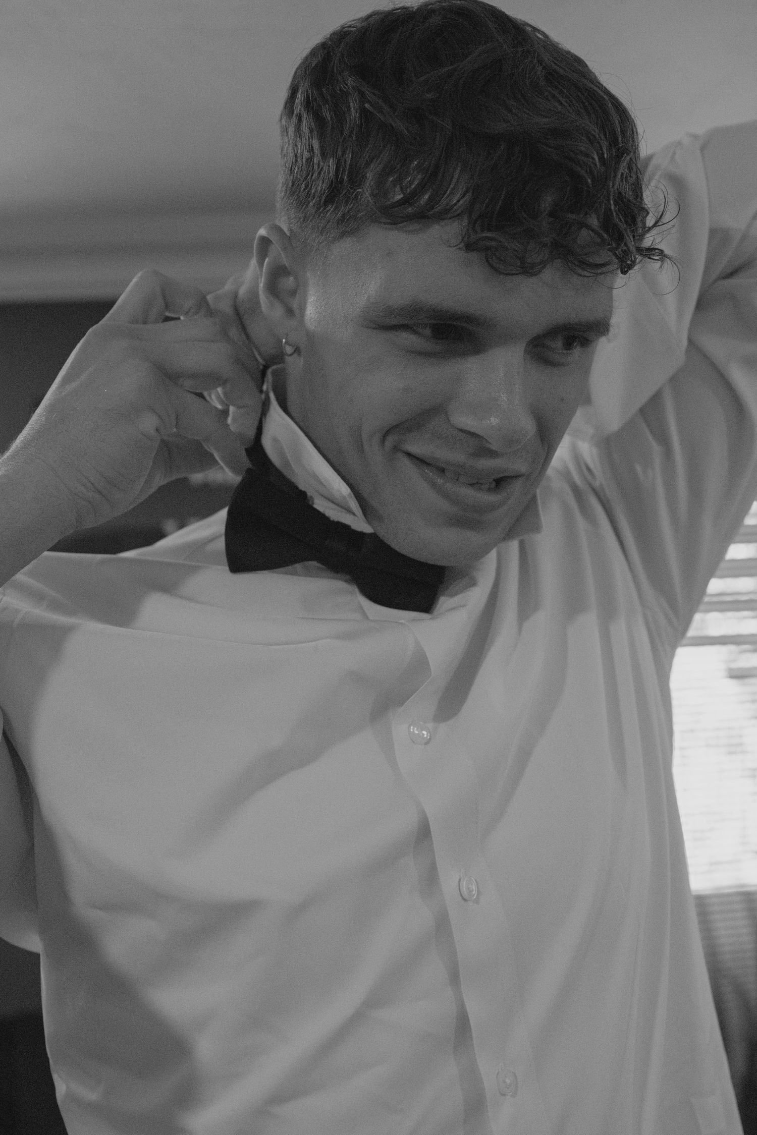 Groom in white dress shirt and black bow tie smiling playfully during wedding day getting ready.