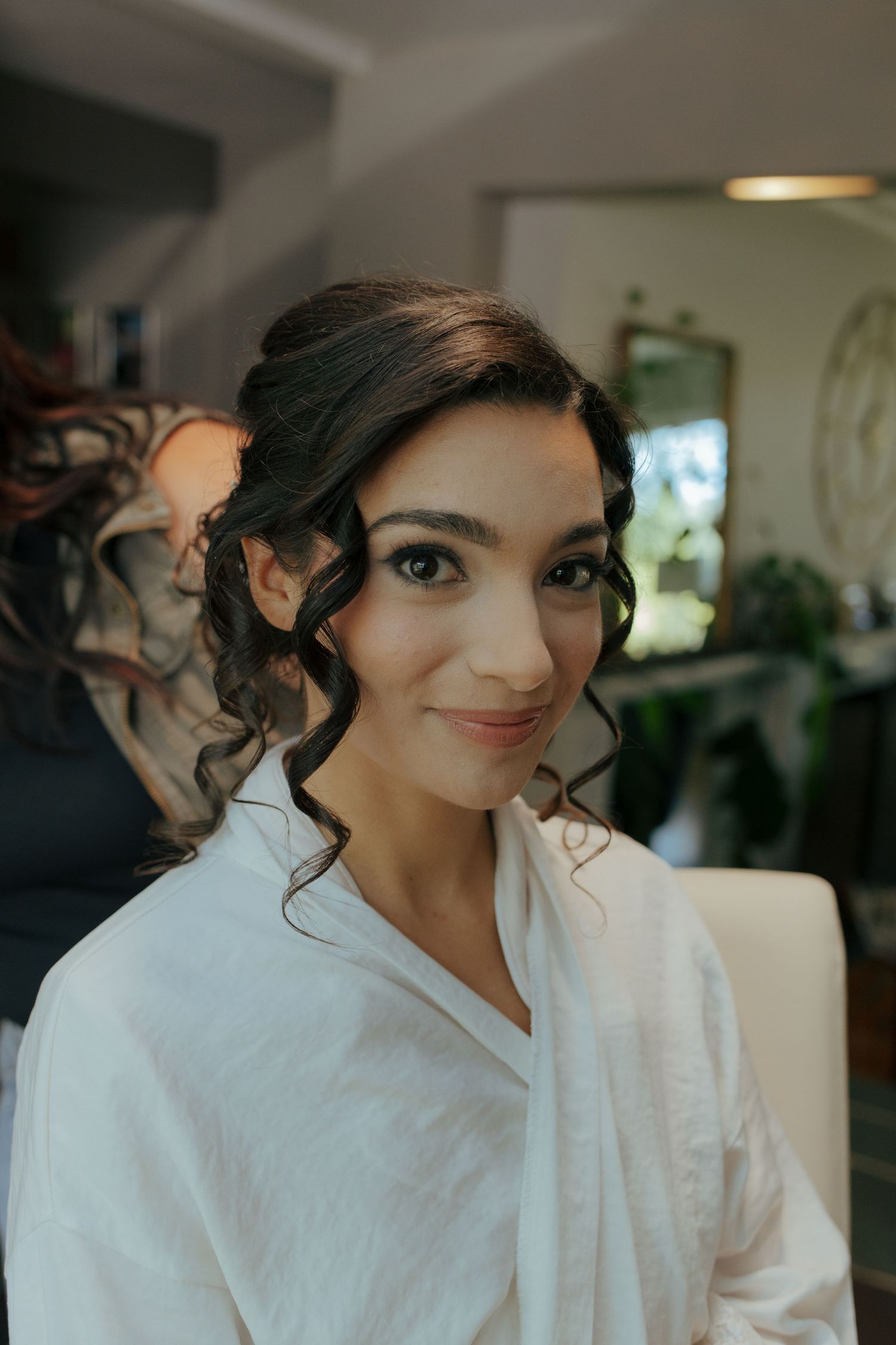 Bride with dark curly updo and natural makeup smiles in white robe during wedding preparations.