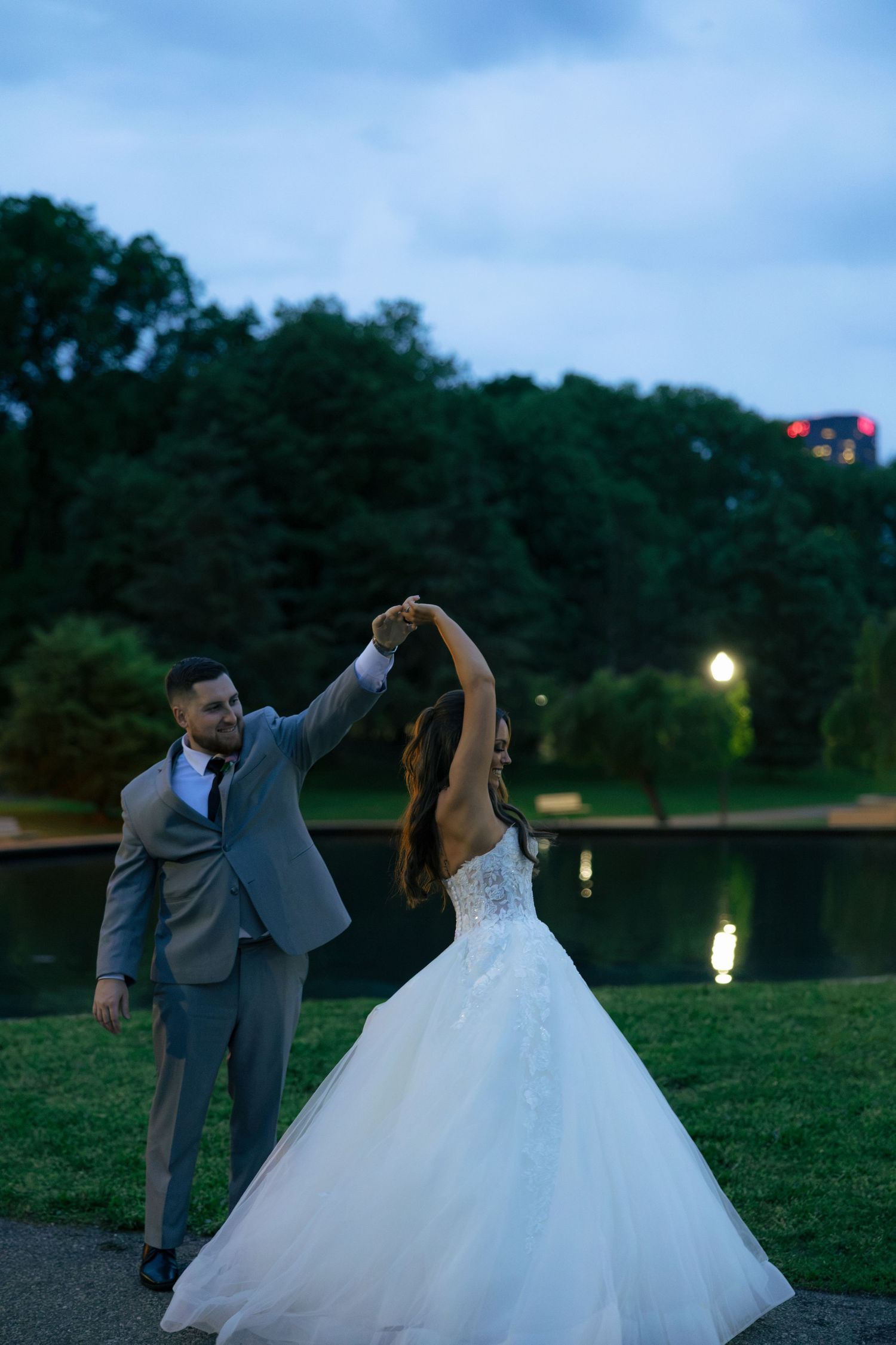Groom in gray suit twirls bride in a lace ballgown near a reflective lake at dusk in a romantic outdoor setting.