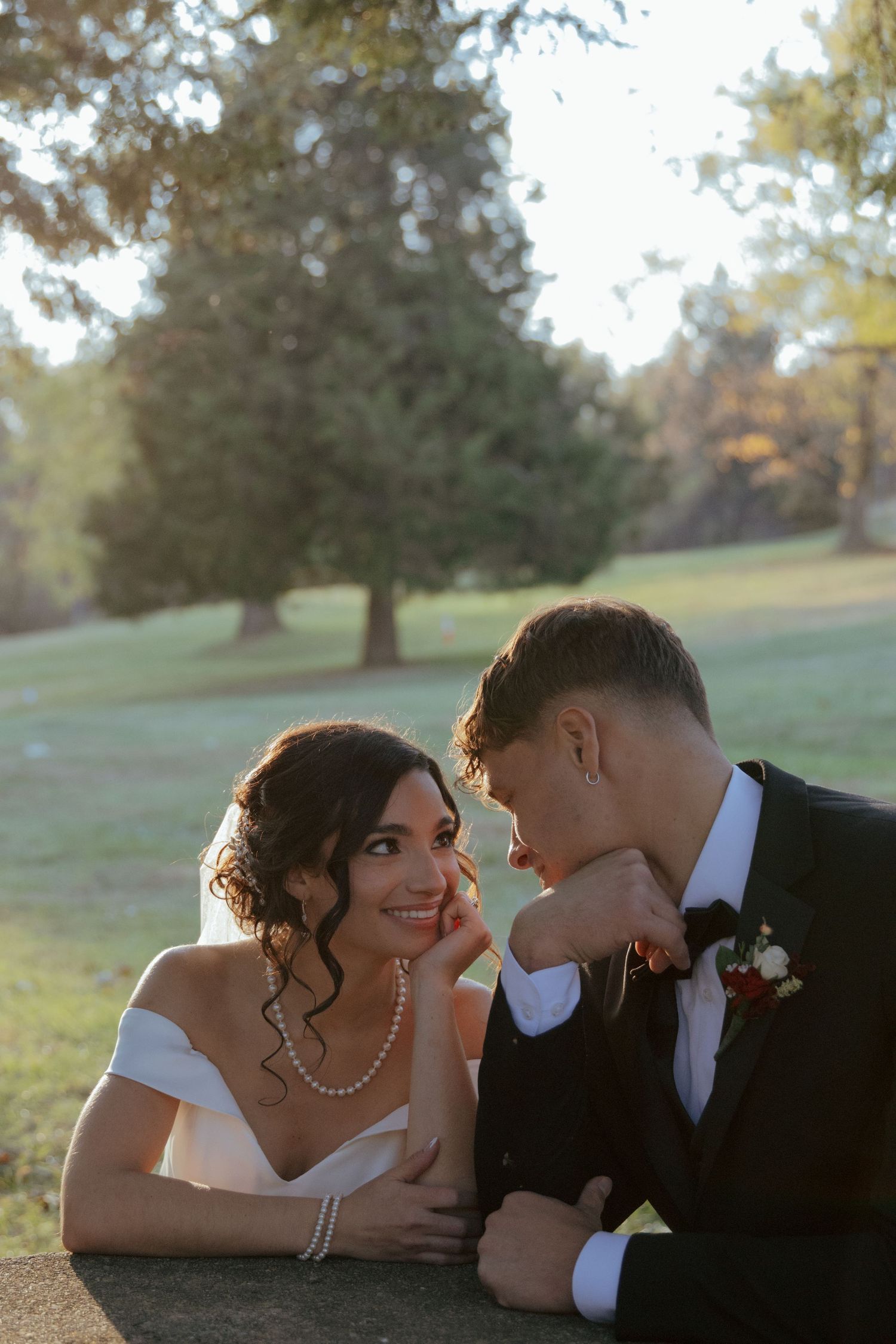 Bride and groom leaning on stone wall gazing at each other lovingly in sunlit park with green trees behind.