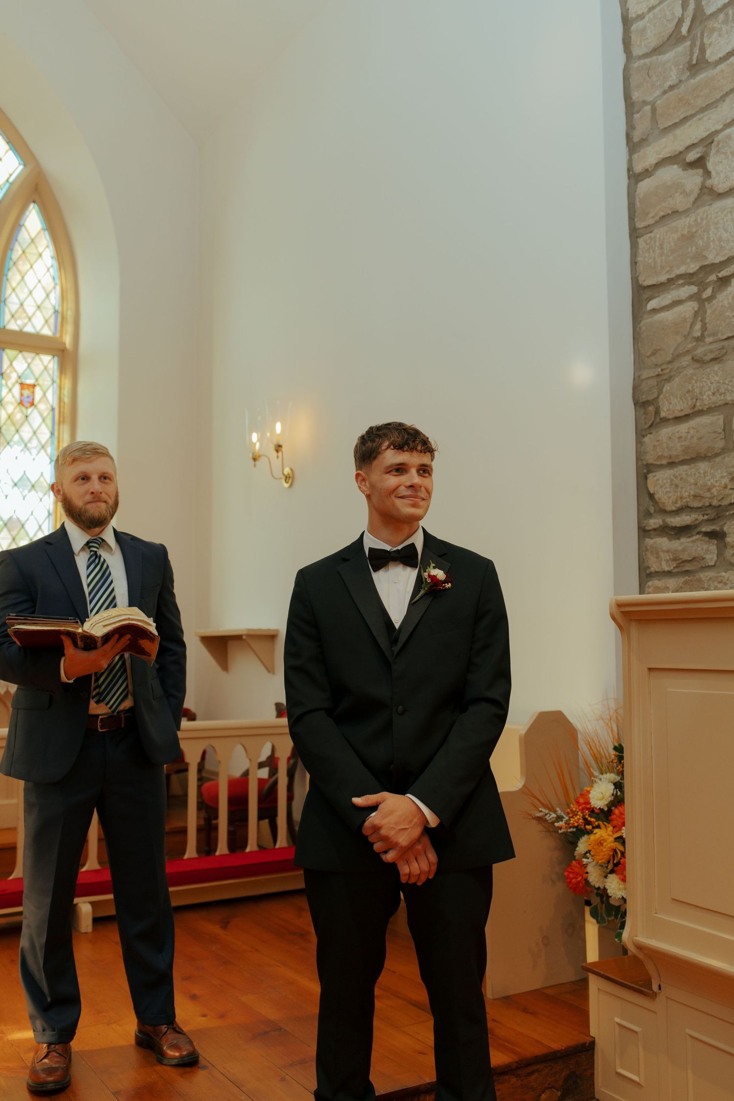 Groom in black tuxedo smiling at altar in stone church, officiant holding Bible beside him.