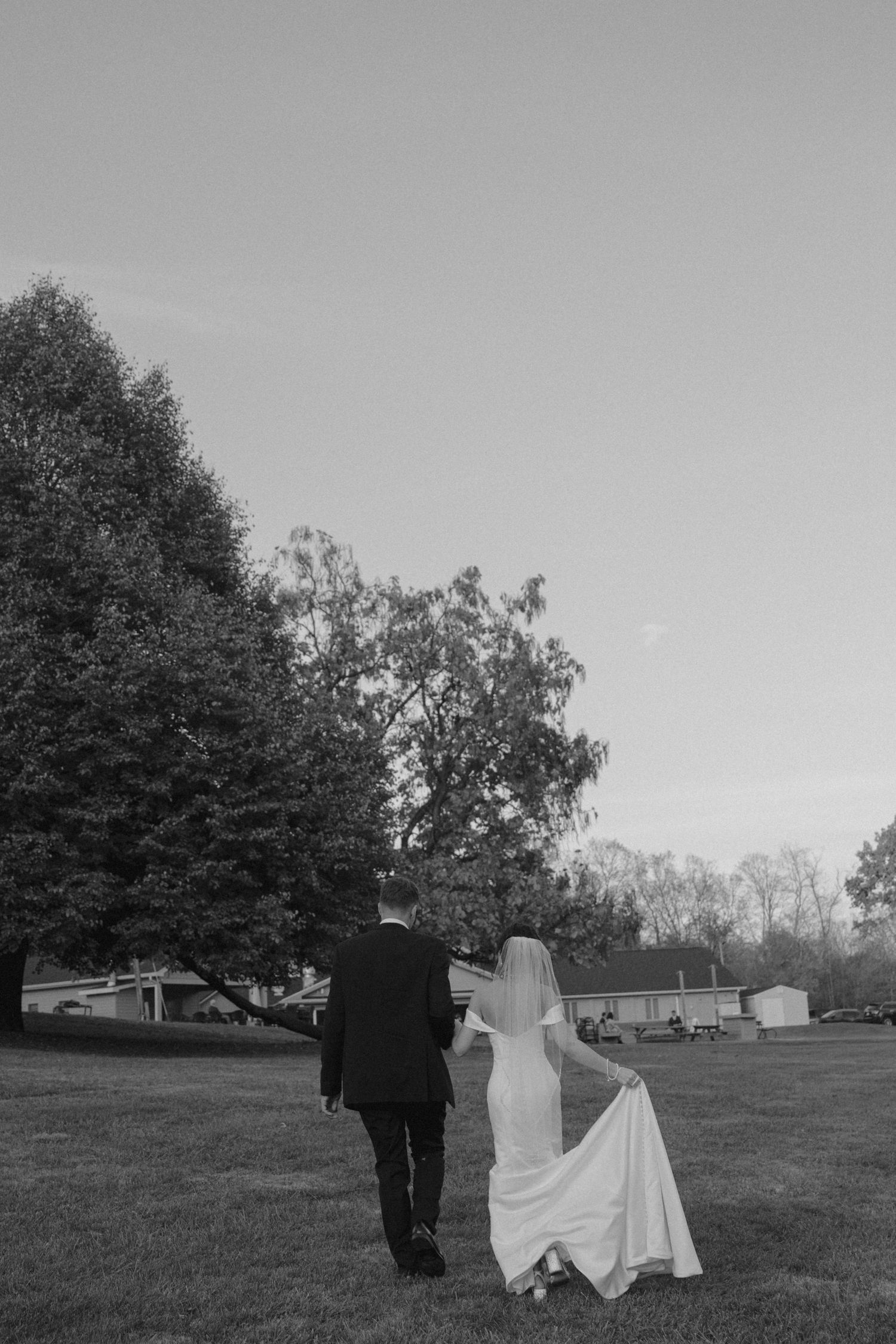 Bride and groom walk across lawn at dusk in black and white.