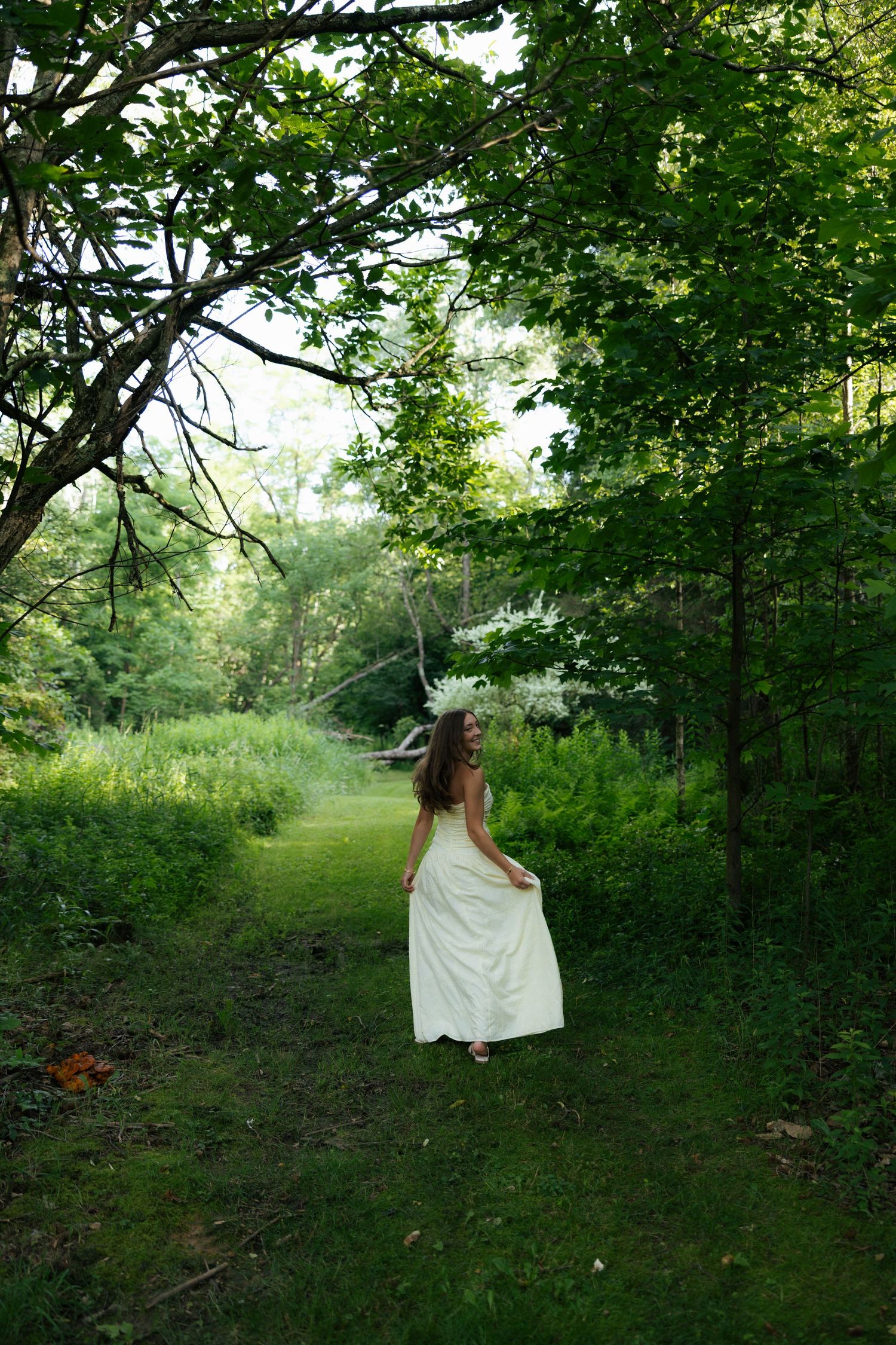 Young woman in white dress standing among tall green trees in a serene woodland garden setting.