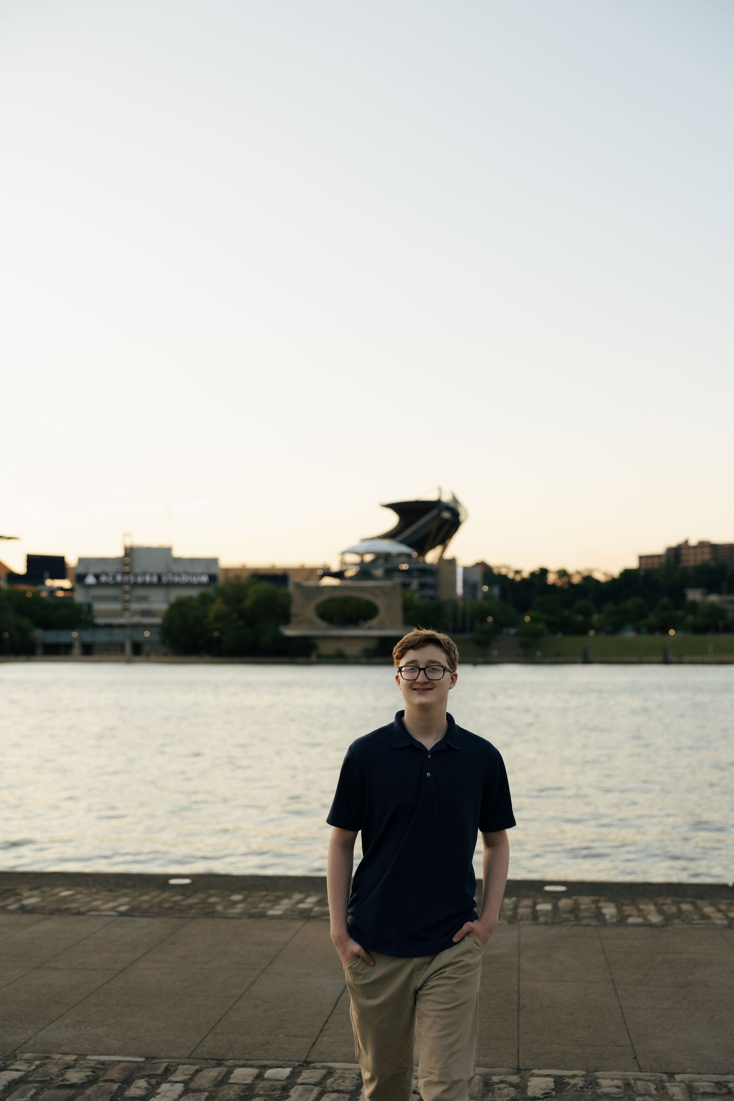 Teen boy in navy polo standing on cobblestone riverfront with Acrisure Stadium visible across the water.