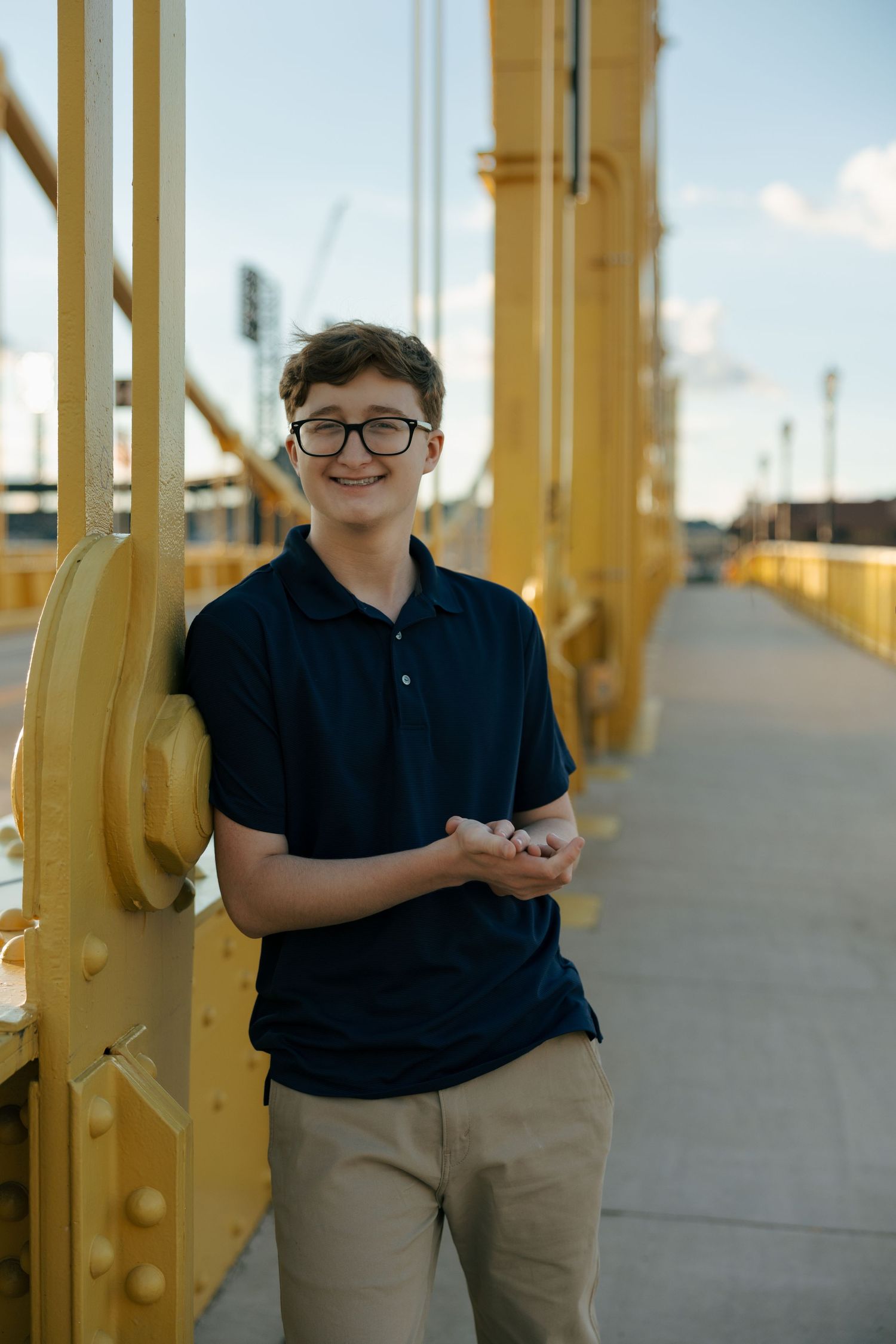 Young man in glasses and navy polo leaning against yellow steel bridge structure, smiling at camera.