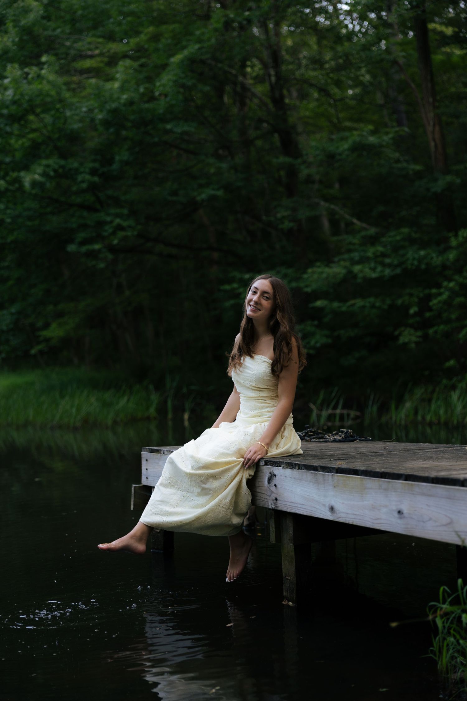 Smiling young woman in cream strapless dress sitting on wooden dock with feet over lake.