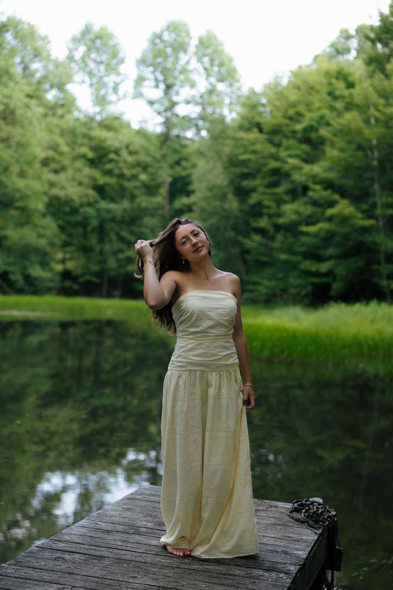 Young woman in cream strapless dress posing on wooden dock by calm lake surrounded by forest.