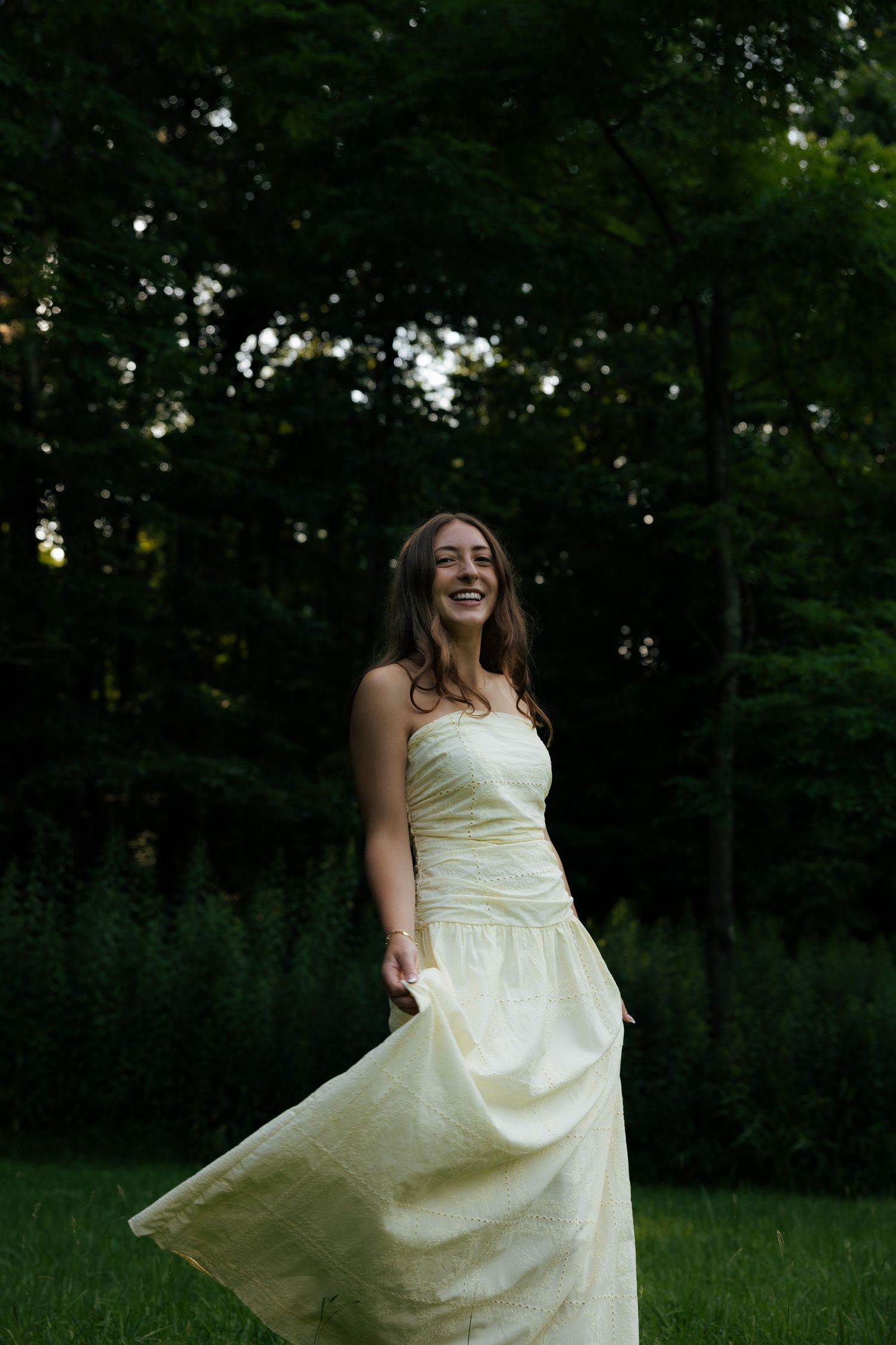 A joyful young woman in a flowing pale yellow strapless gown twirling outdoors in a lush green garden.