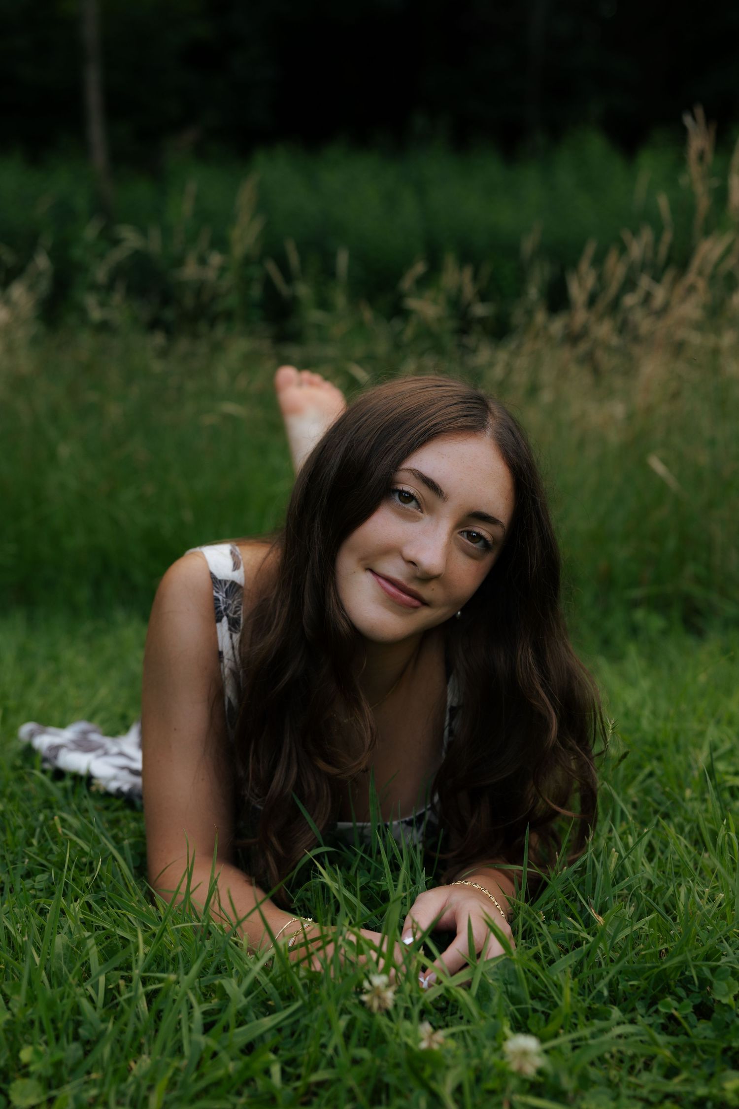 Young woman with long brown hair lying on green grass, smiling at camera in a floral dress outdoors.