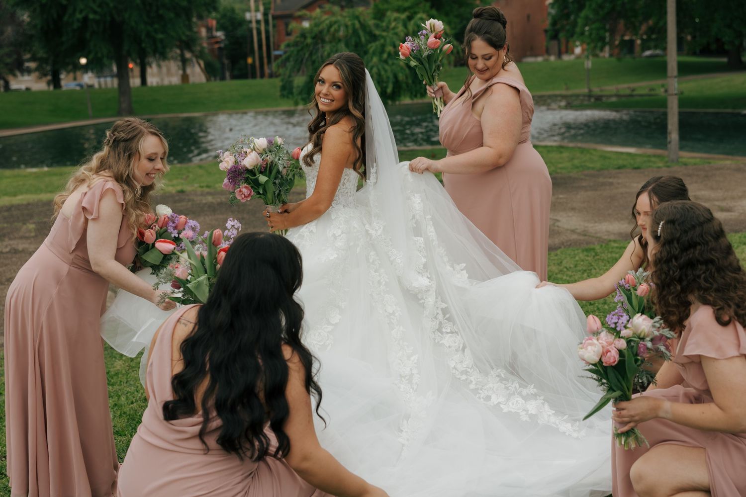Smiling bride in lace gown surrounded by bridesmaids in blush dresses holding colorful bouquets outdoors.