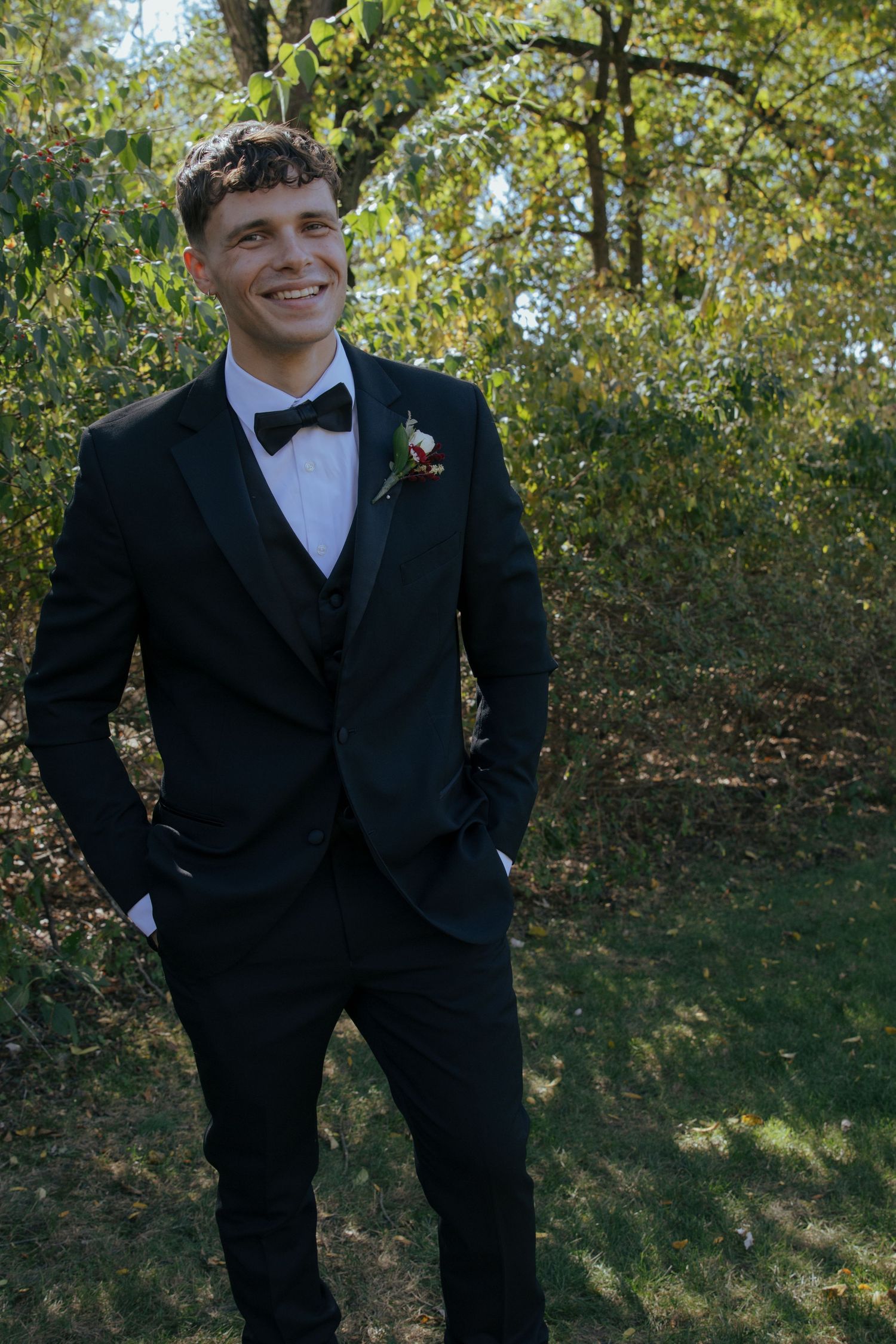 Smiling groom in black three-piece tuxedo with bow tie and boutonniere posing outdoors among green trees.