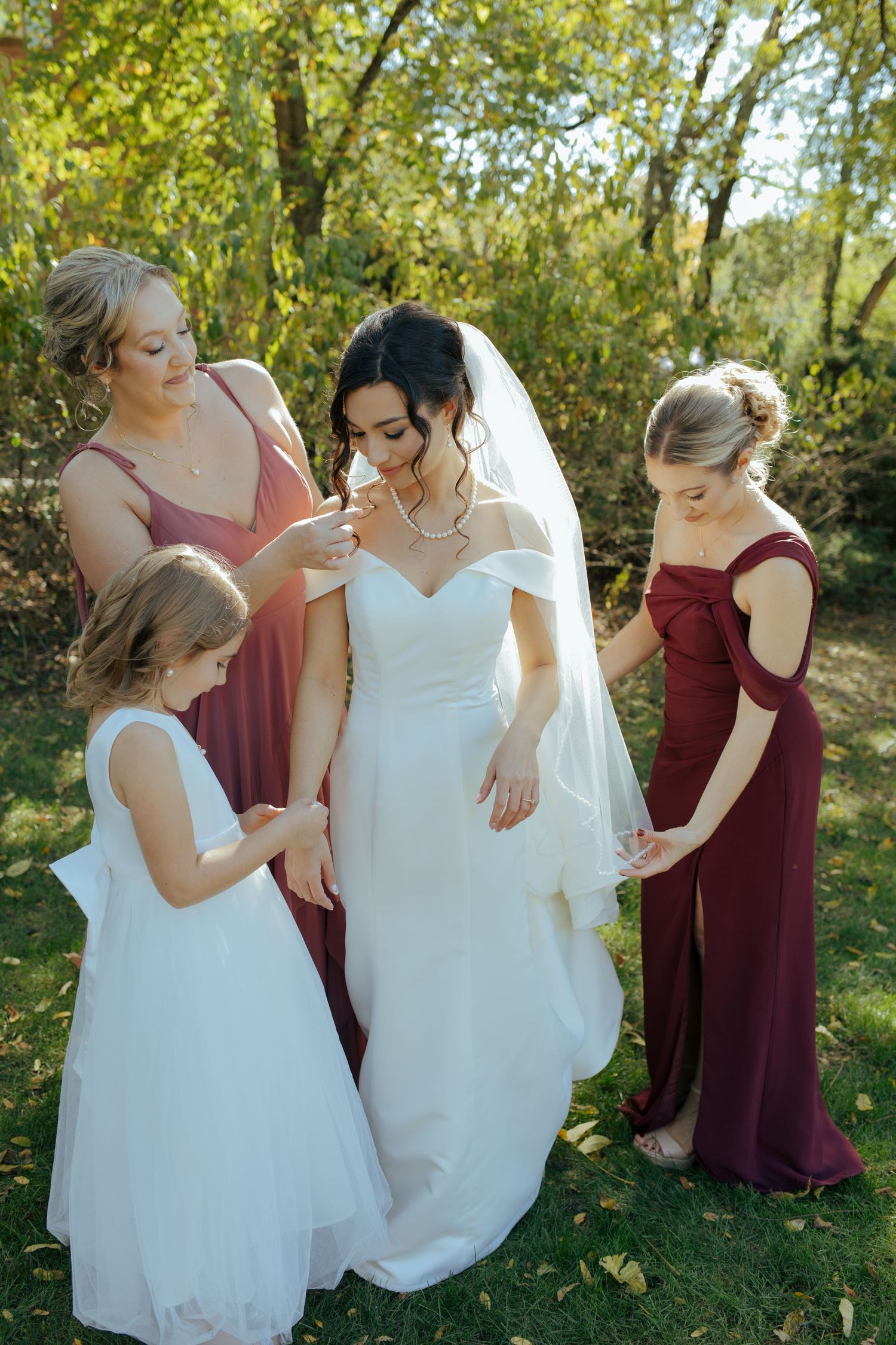 Bride in white gown with veil surrounded by bridesmaids in burgundy dresses and flower girl outdoors.