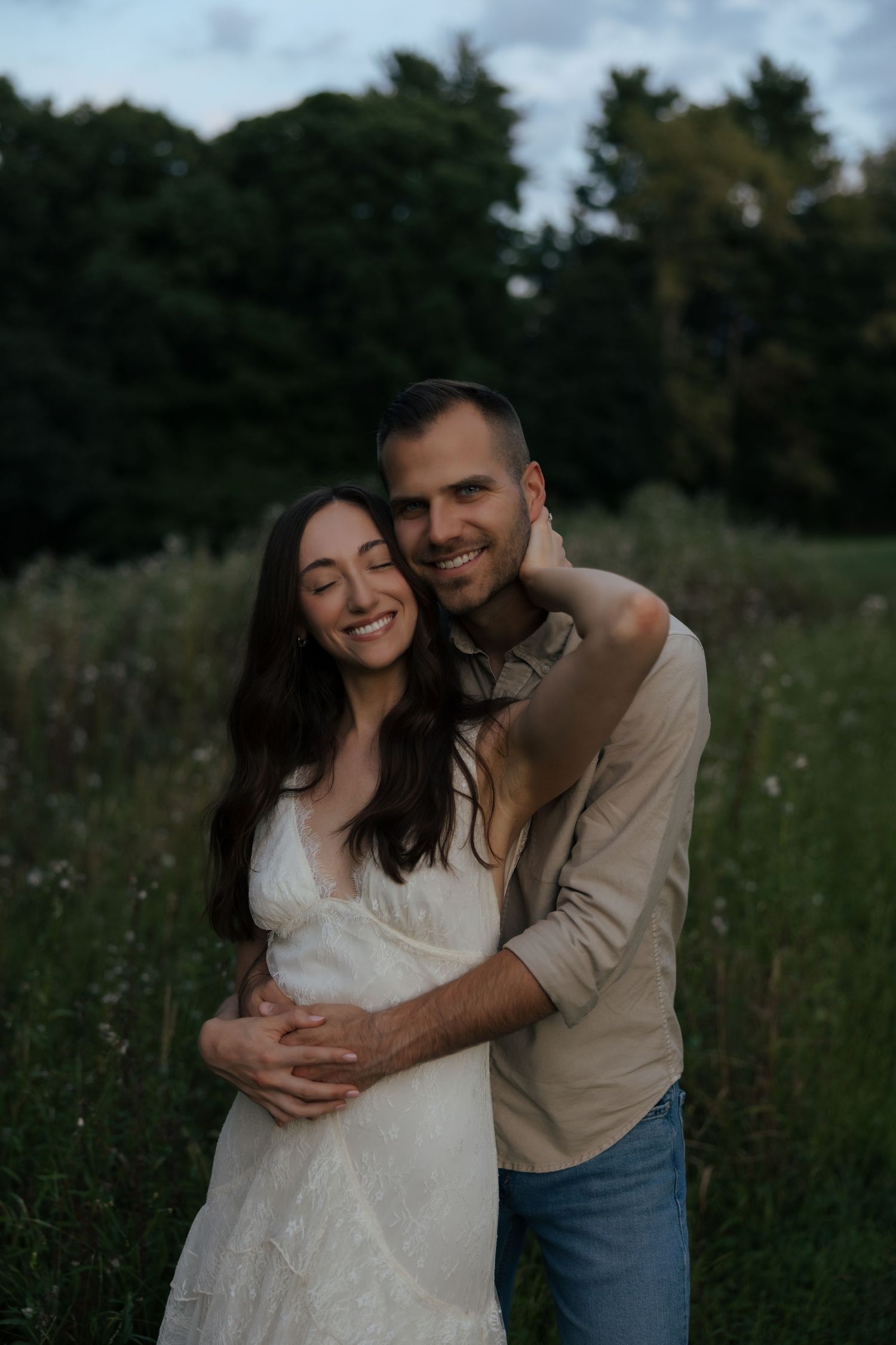 Couple embracing in a field at dusk, woman in white dress smiling with eyes closed, man hugging her from behind.