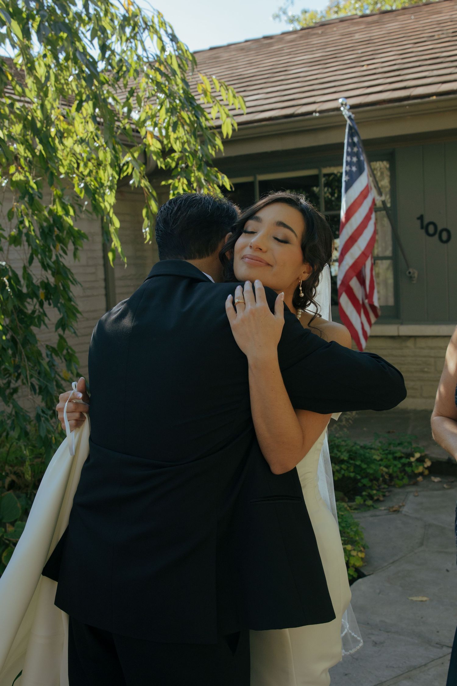 Couple embracing outdoors near a brick house with an American flag, woman smiling with eyes closed in formal attire.