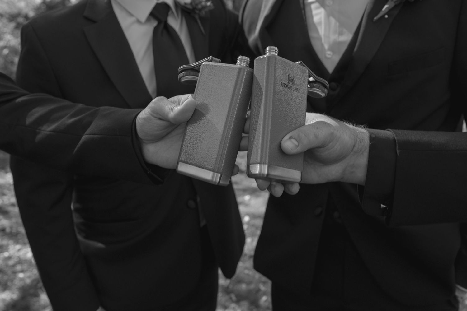 Two men in suits toasting with metal flasks in a black and white wedding photo.