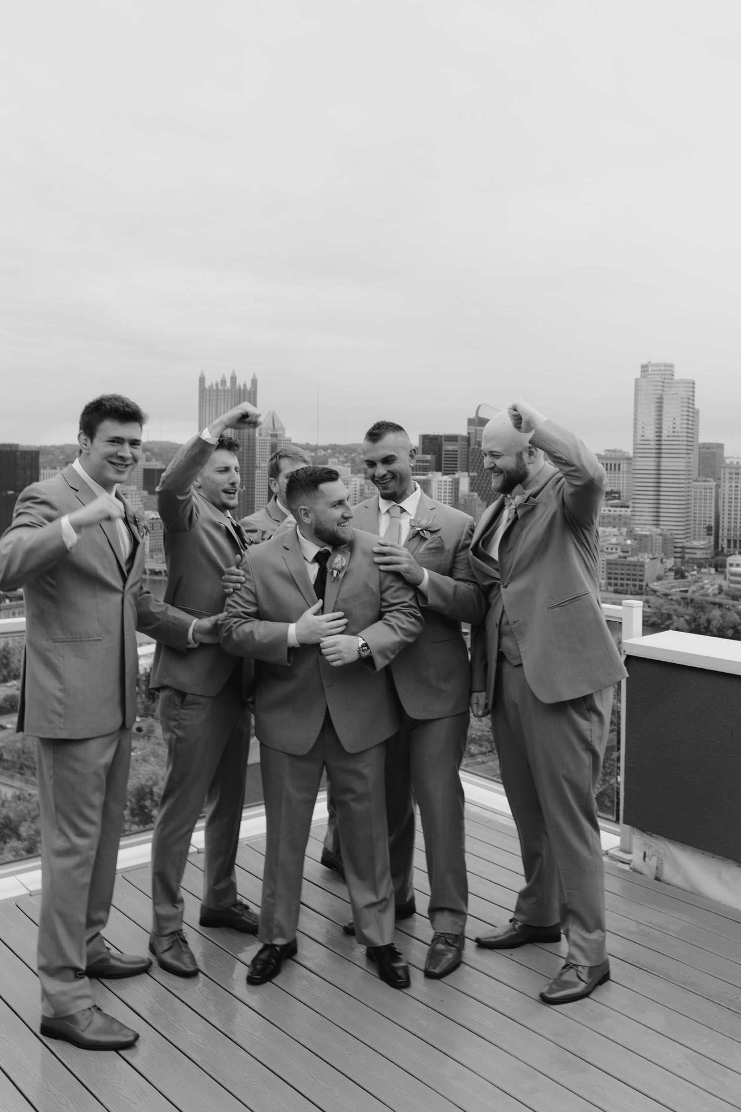 Five groomsmen in gray suits celebrate on a rooftop with Pittsburgh city skyline in the background.