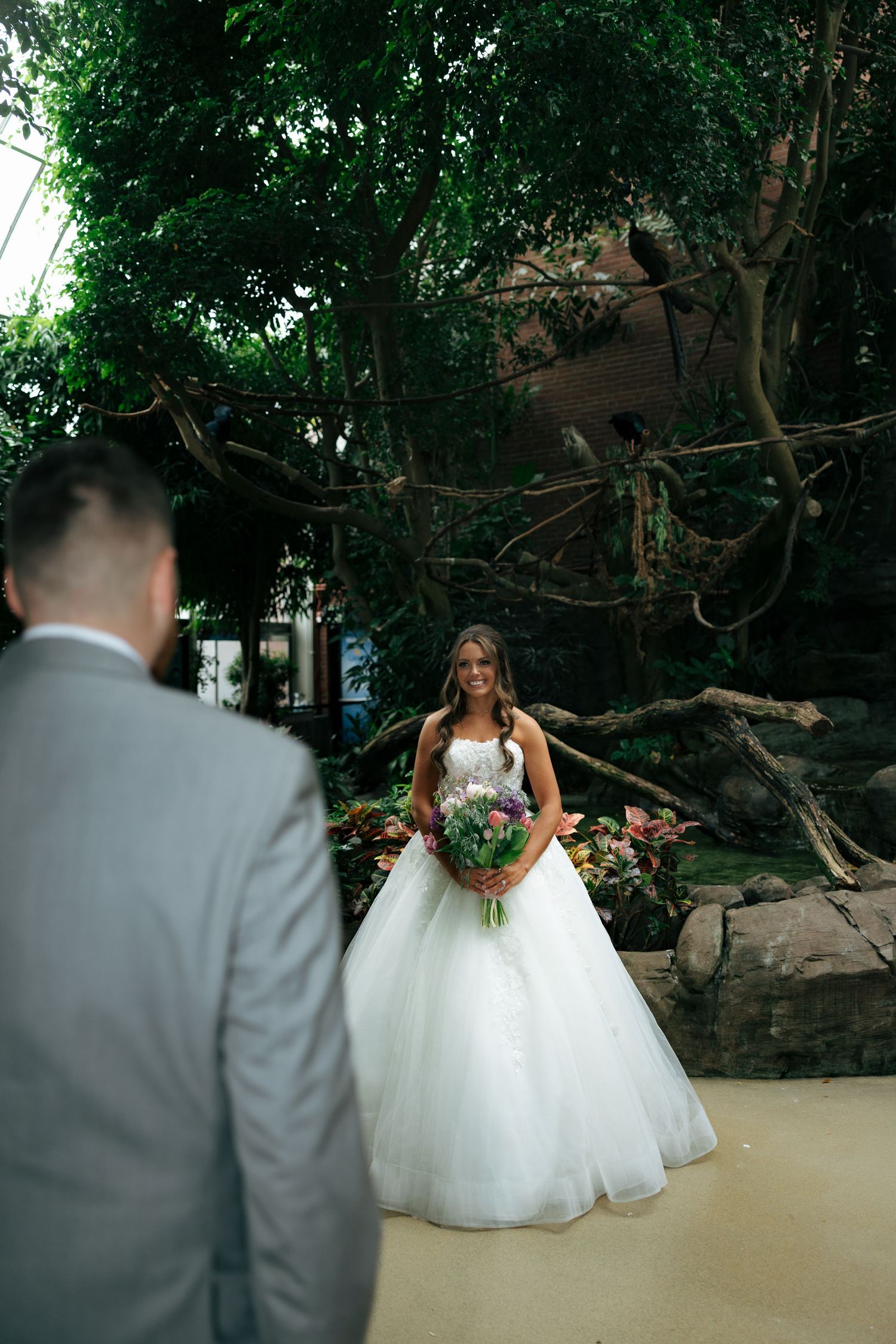 Smiling bride in a ball gown holds bouquet during first look with groom in botanical garden setting.