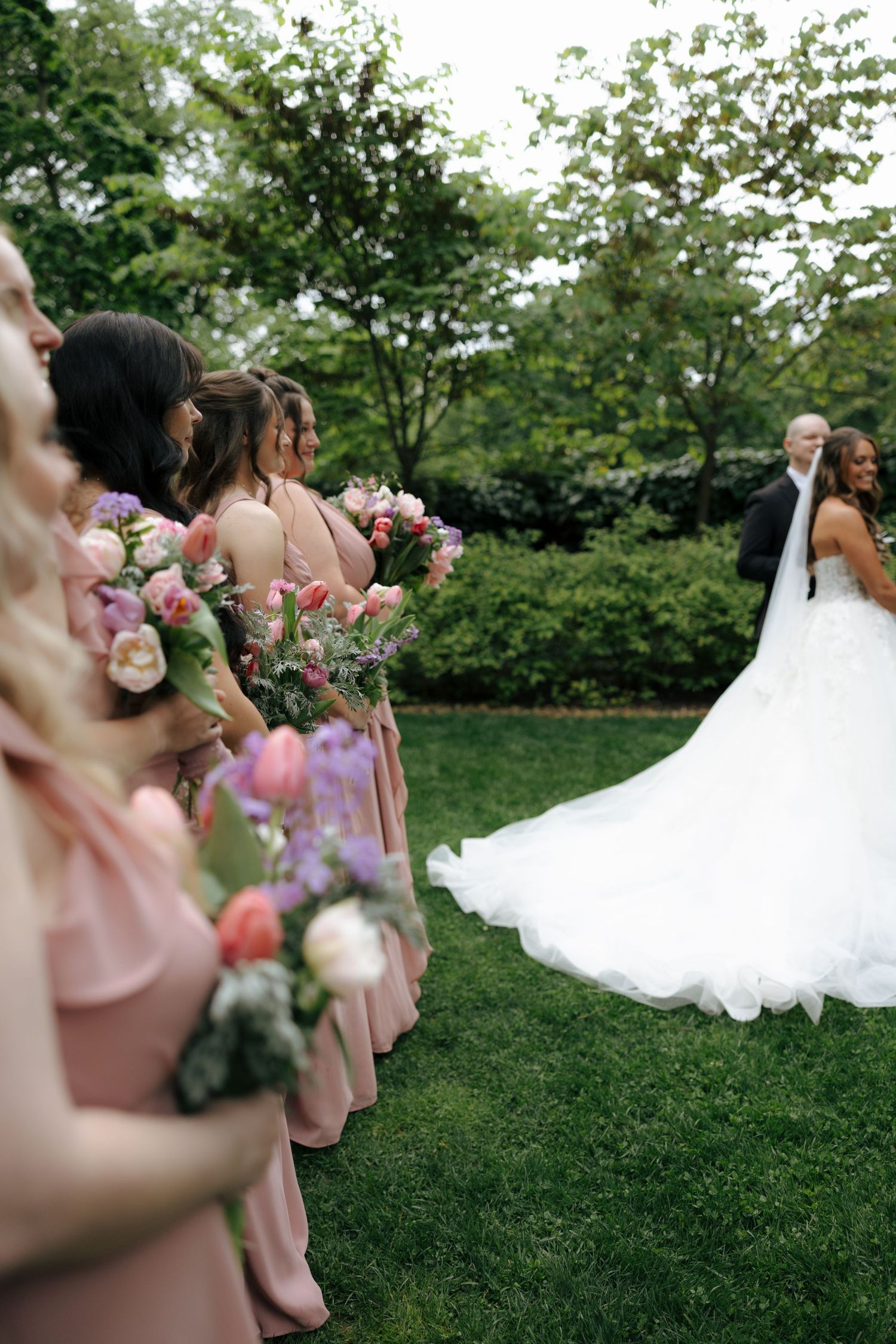 Bridesmaids in blush pink dresses holding colorful bouquets stand at outdoor garden wedding ceremony.
