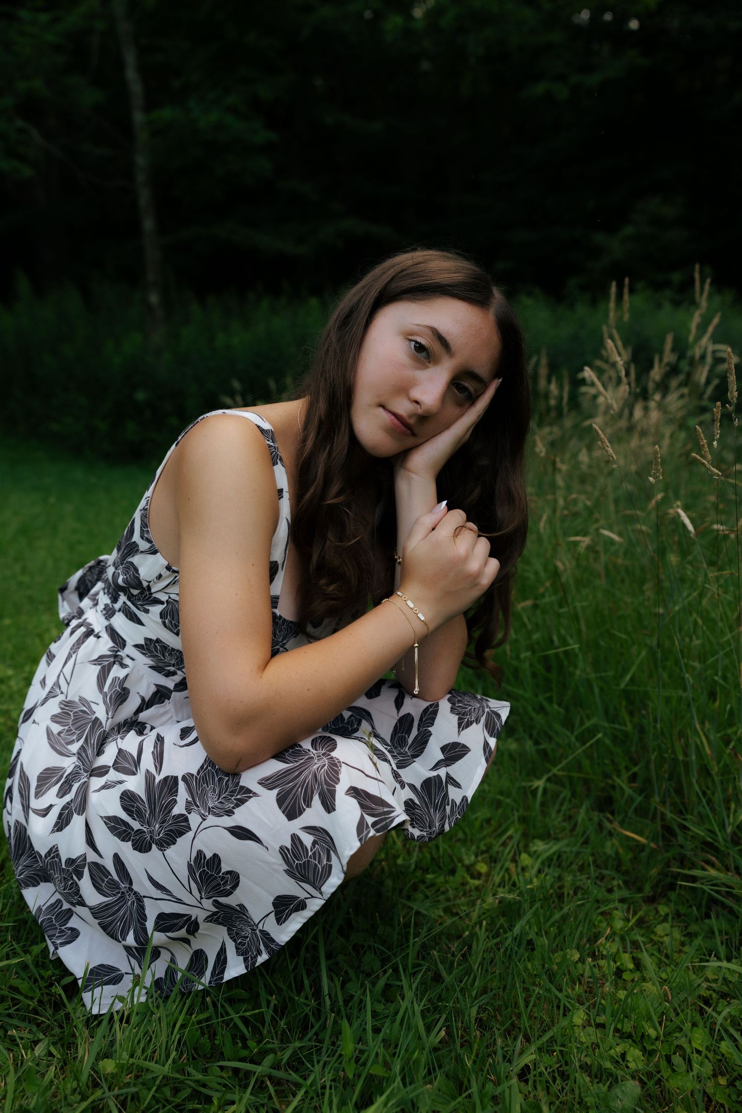 Young woman in black and white floral dress sitting on grass resting chin on hand outdoors.