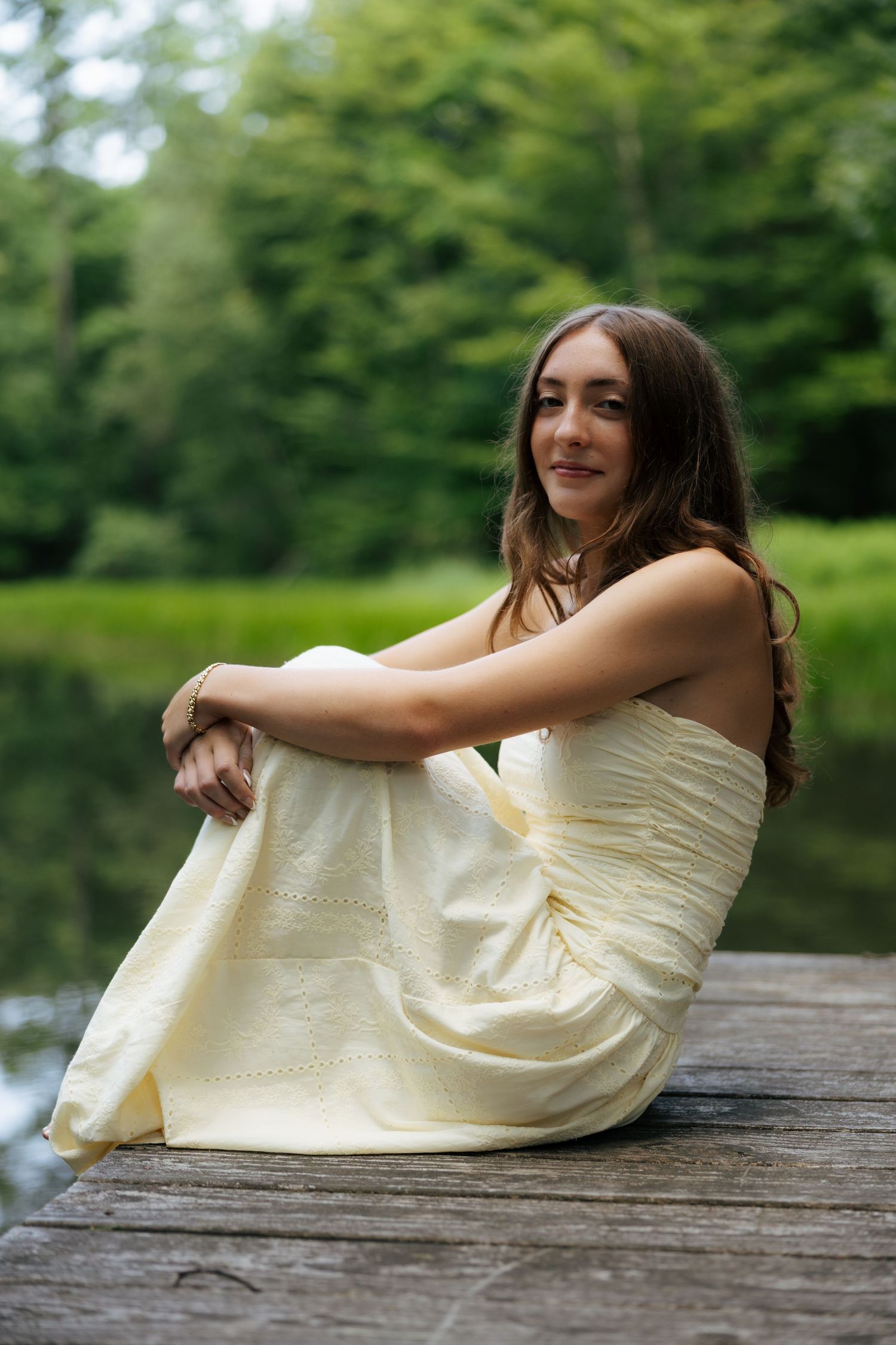 Young woman in cream dress sitting on wooden dock by lake, surrounded by green trees.