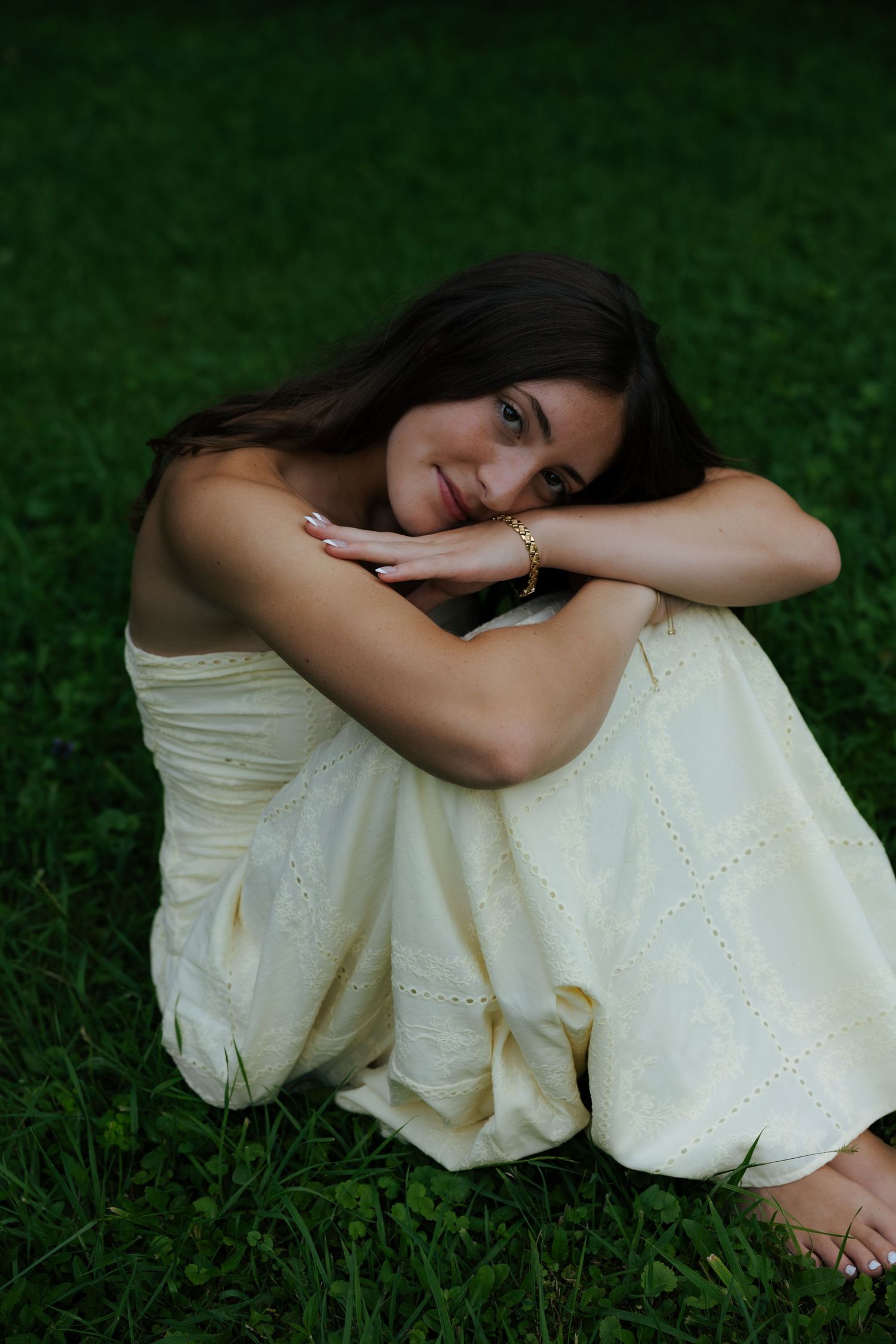 Brunette woman in cream dress sitting barefoot on green grass with knees drawn up smiling.