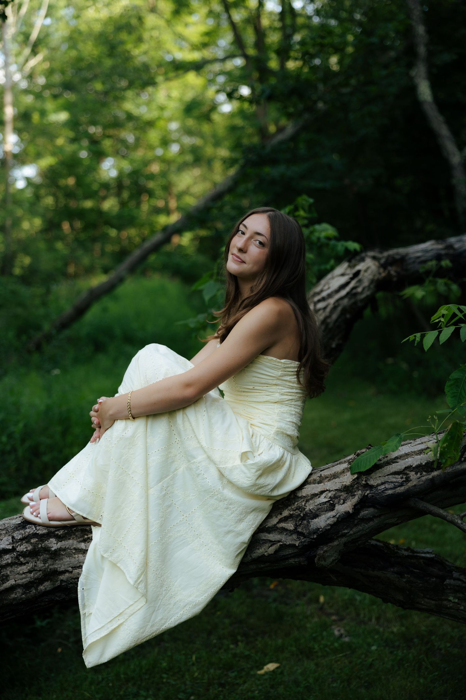 Young woman in strapless cream gown sitting gracefully on fallen log in forest.
