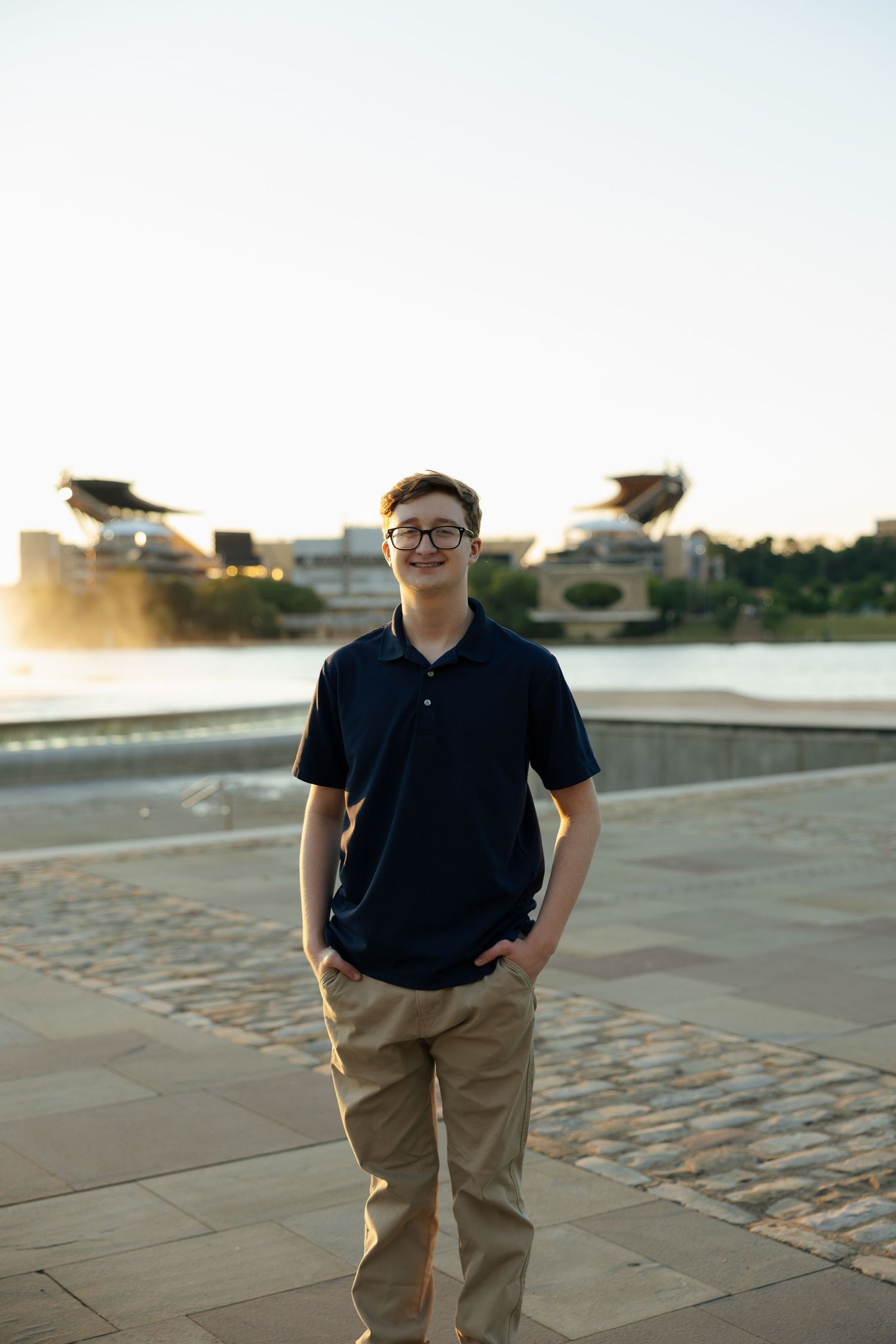 Smiling teenage boy in navy polo and khakis standing on riverside plaza with stadium in background.