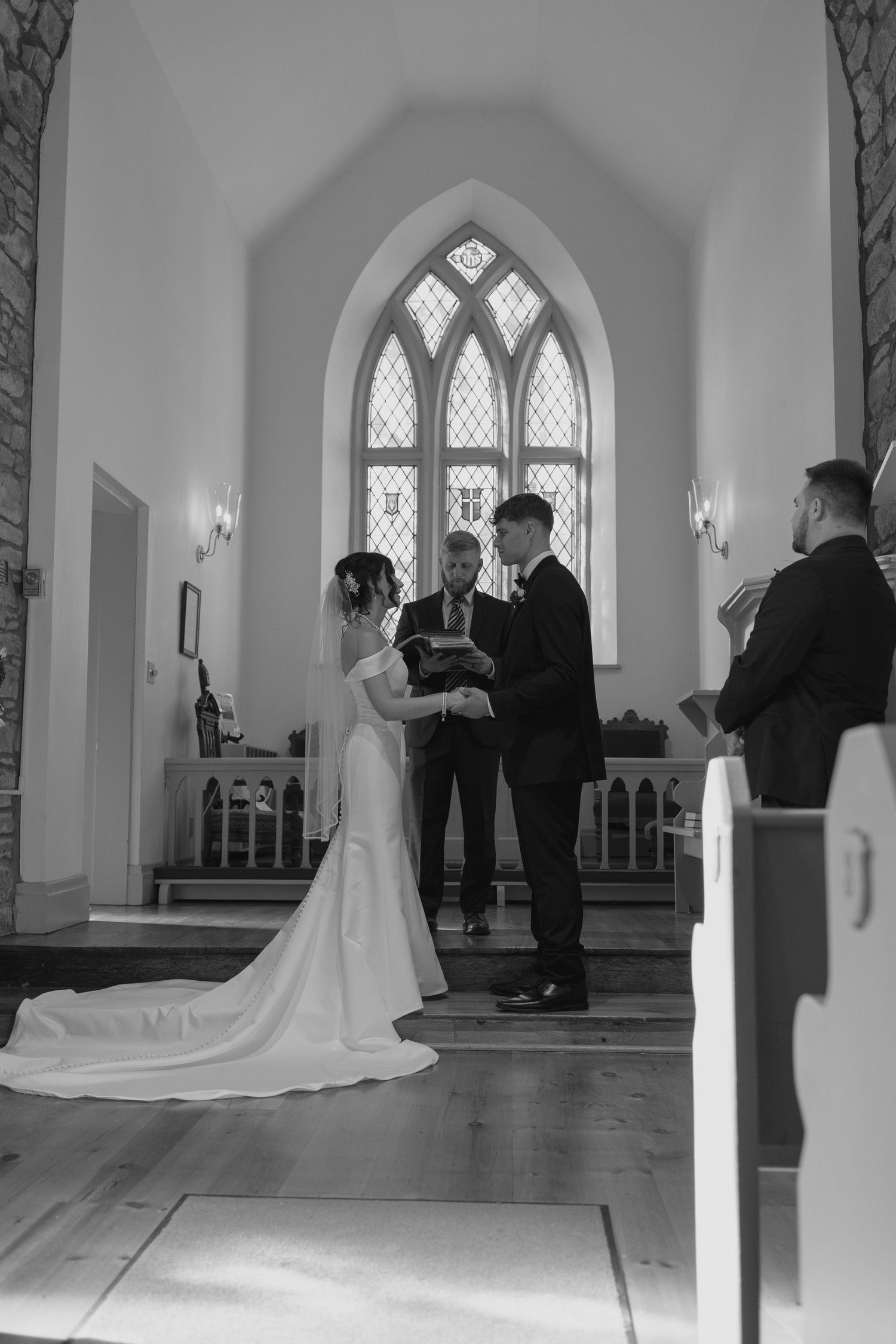 Bride and groom exchange vows at the altar of a charming chapel with arched stained glass windows.