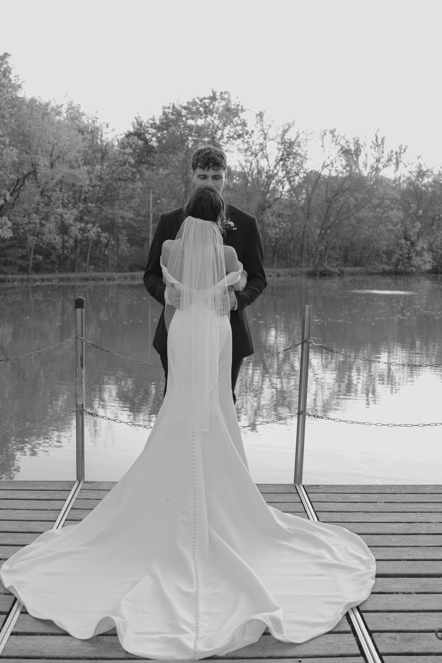Black and white photo of bride and groom standing on dock overlooking serene lake.