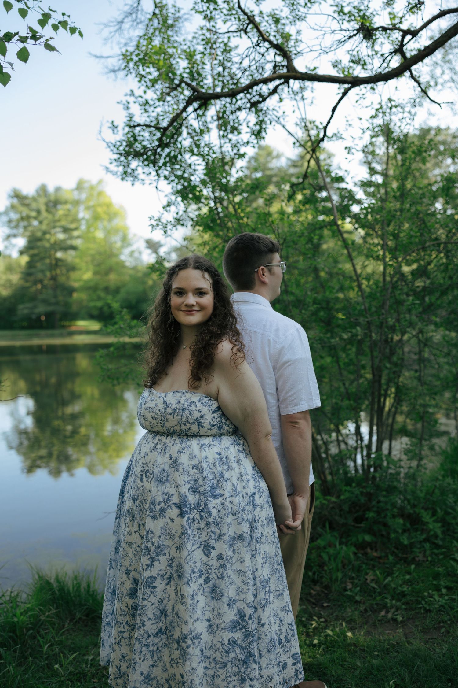 Couple holding hands by a serene pond in a lush green park, woman in floral dress smiling at camera.