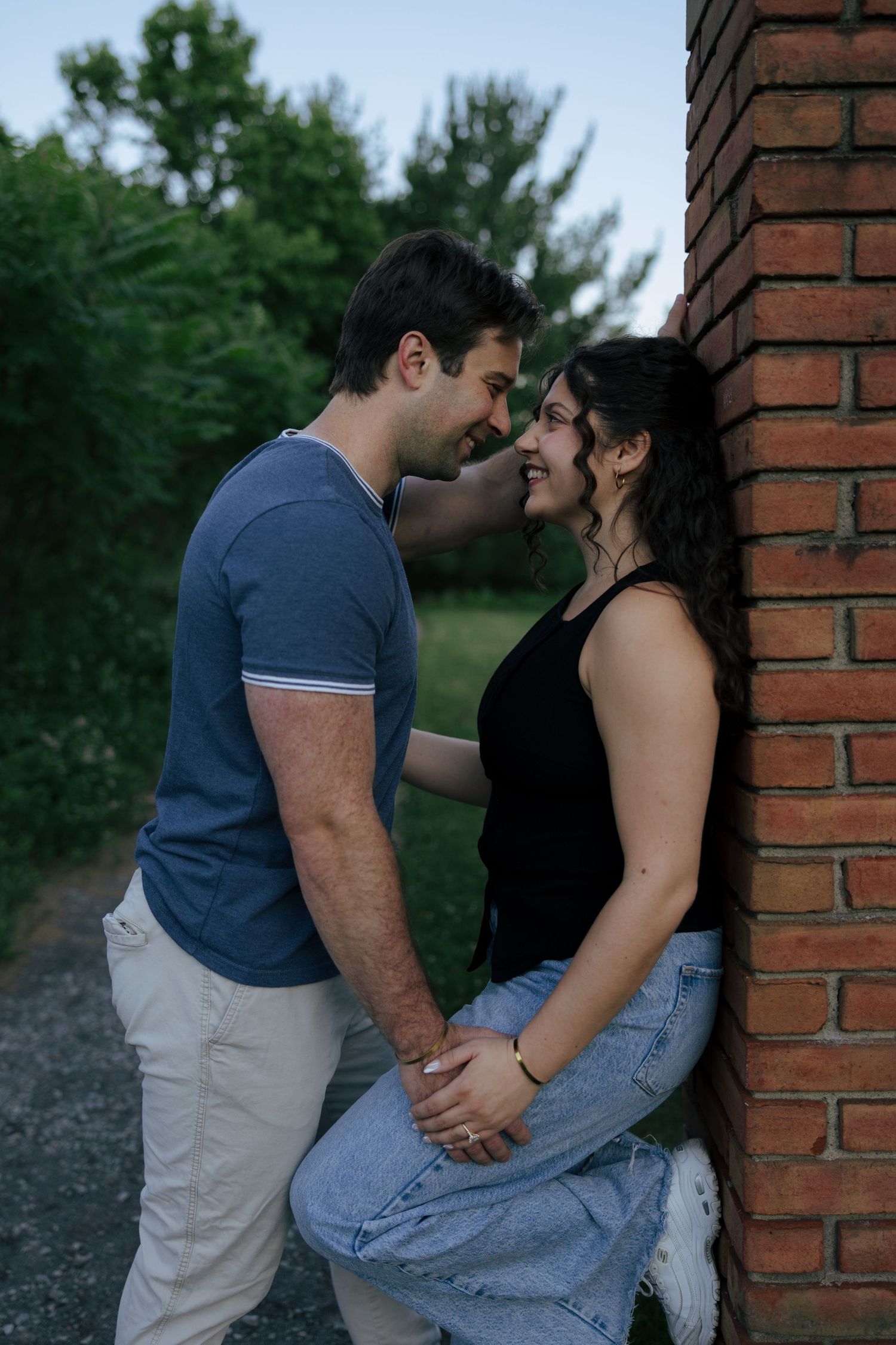 Romantic couple sharing an intimate moment leaning against a brick wall outdoors, surrounded by lush green trees.