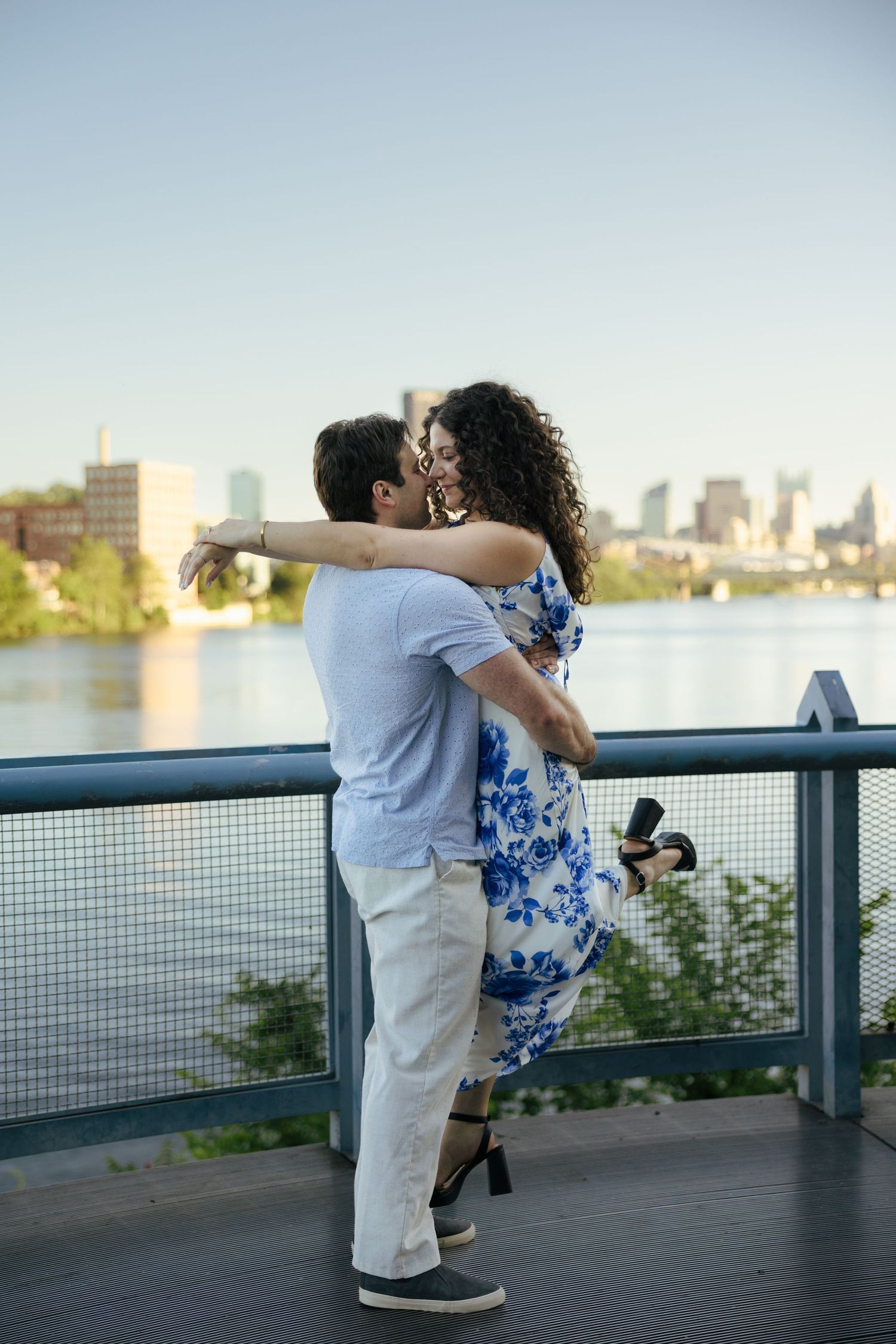 A couple shares a kiss on a bridge railing with a city skyline and river visible behind them.