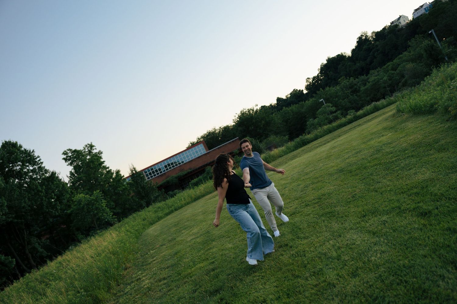 Couple running and laughing together across a green grassy hill at sunset near a covered bridge.