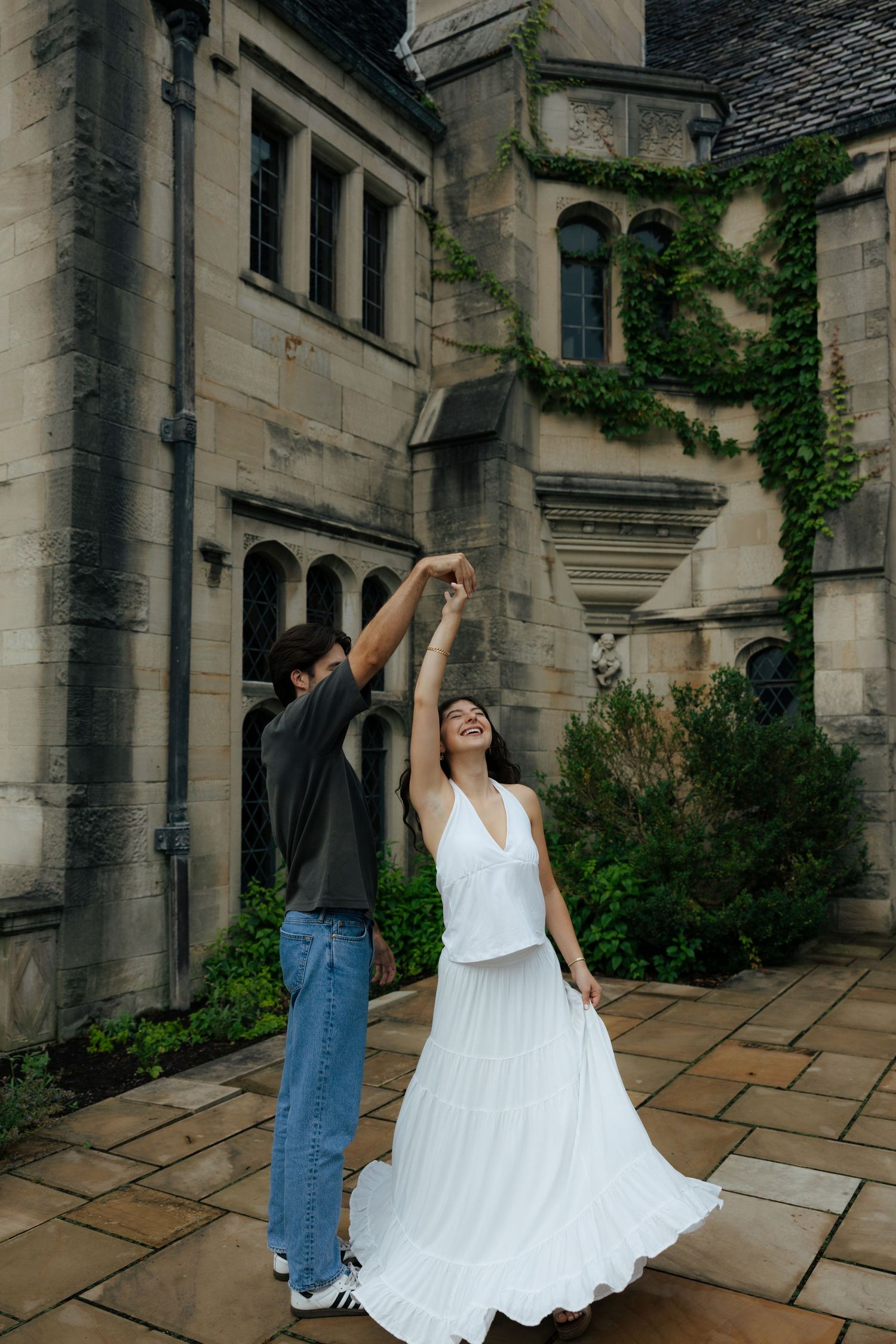 Couple dancing joyfully in courtyard of ivy-covered stone castle building, woman in white dress spinning.