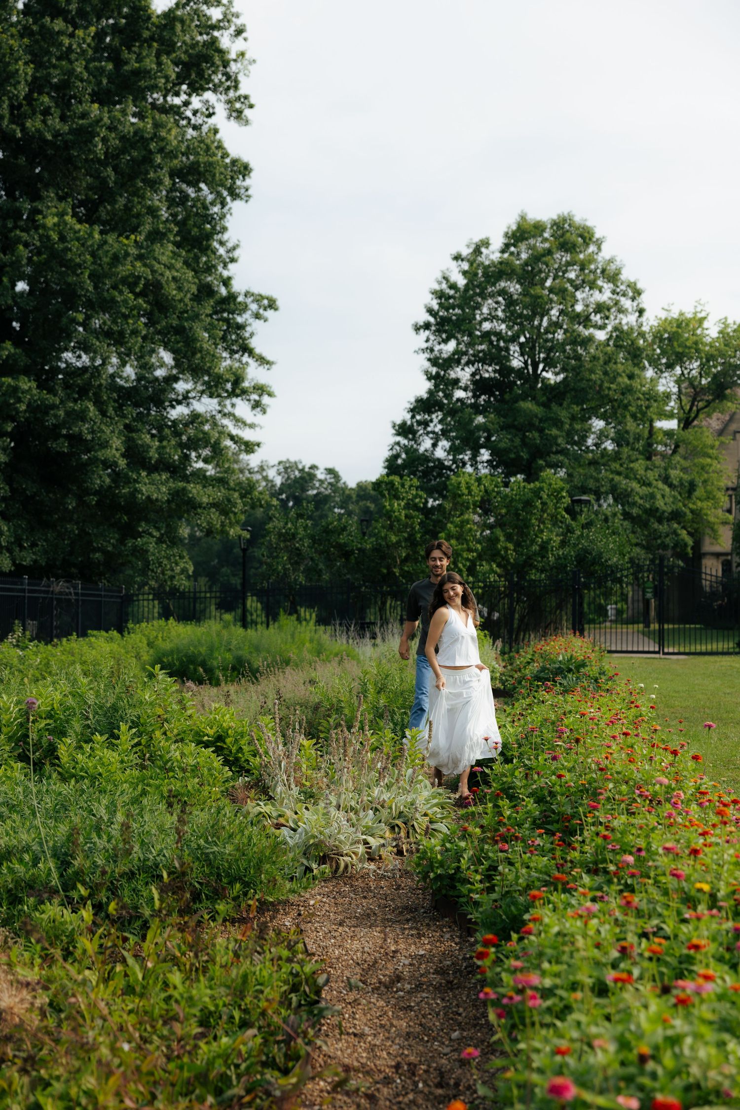 A couple walks together along a garden path lined with colorful flowers and tall trees.