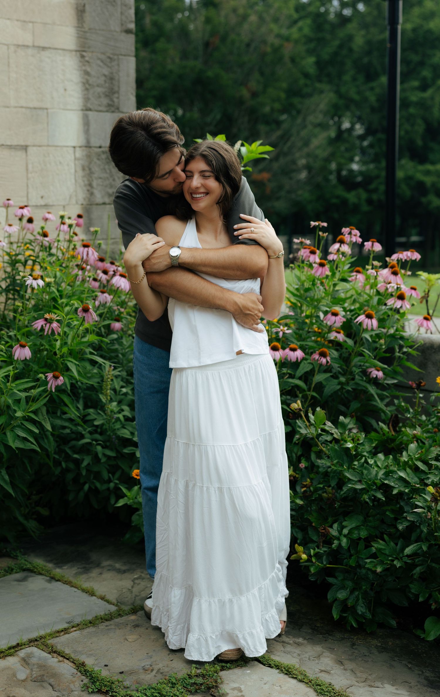 A happy couple embraces among pink wildflowers, the woman in a white maxi dress smiling warmly.