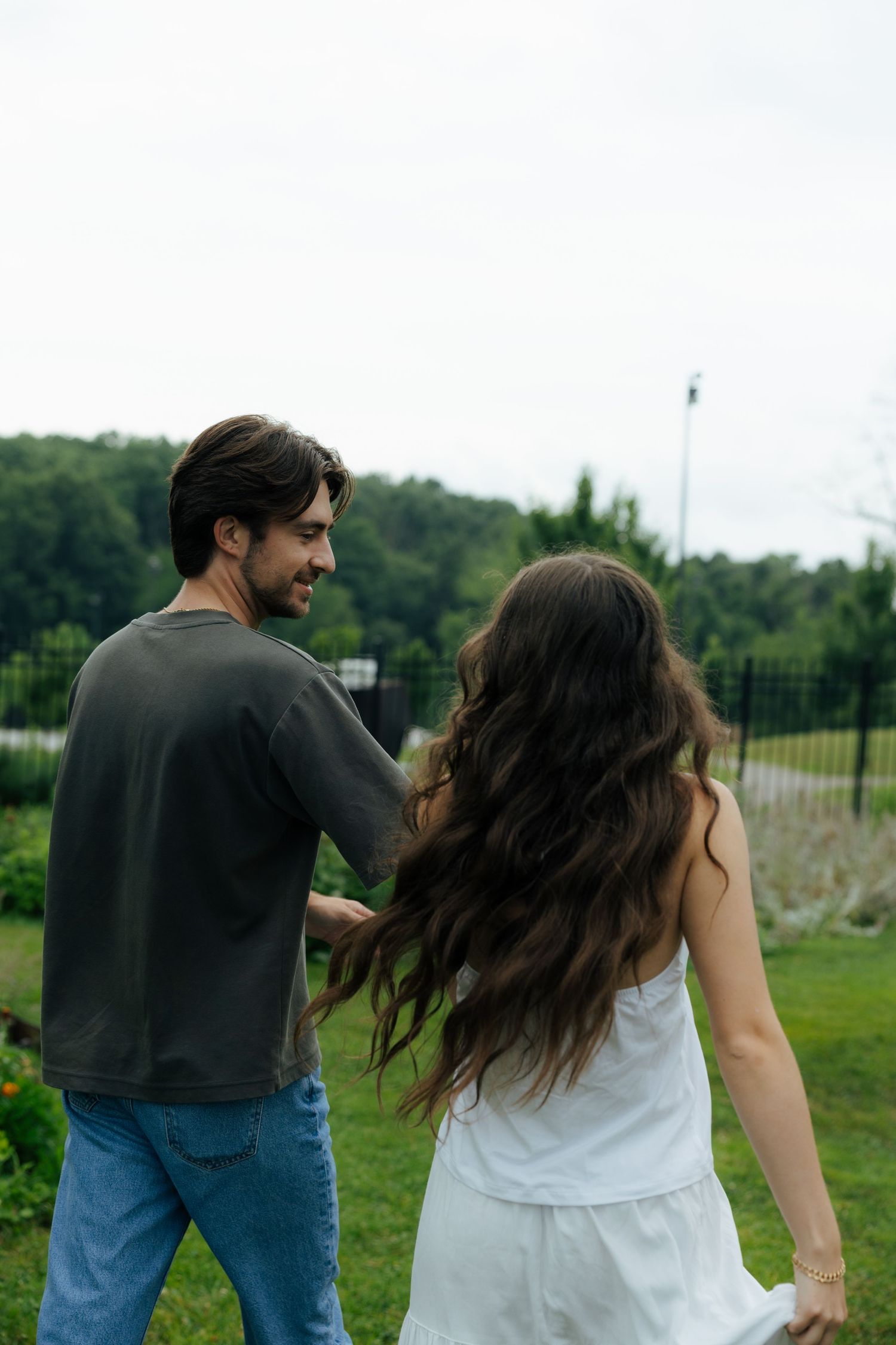 A smiling couple walking hand in hand through a green garden, man in gray shirt and jeans, woman in white dress.